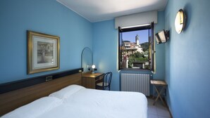 A hotel room with a bed, a desk, a chair, a radiator, a window with a view of buildings, and a framed picture on the wall. - Albergo Sala (Valbrona)