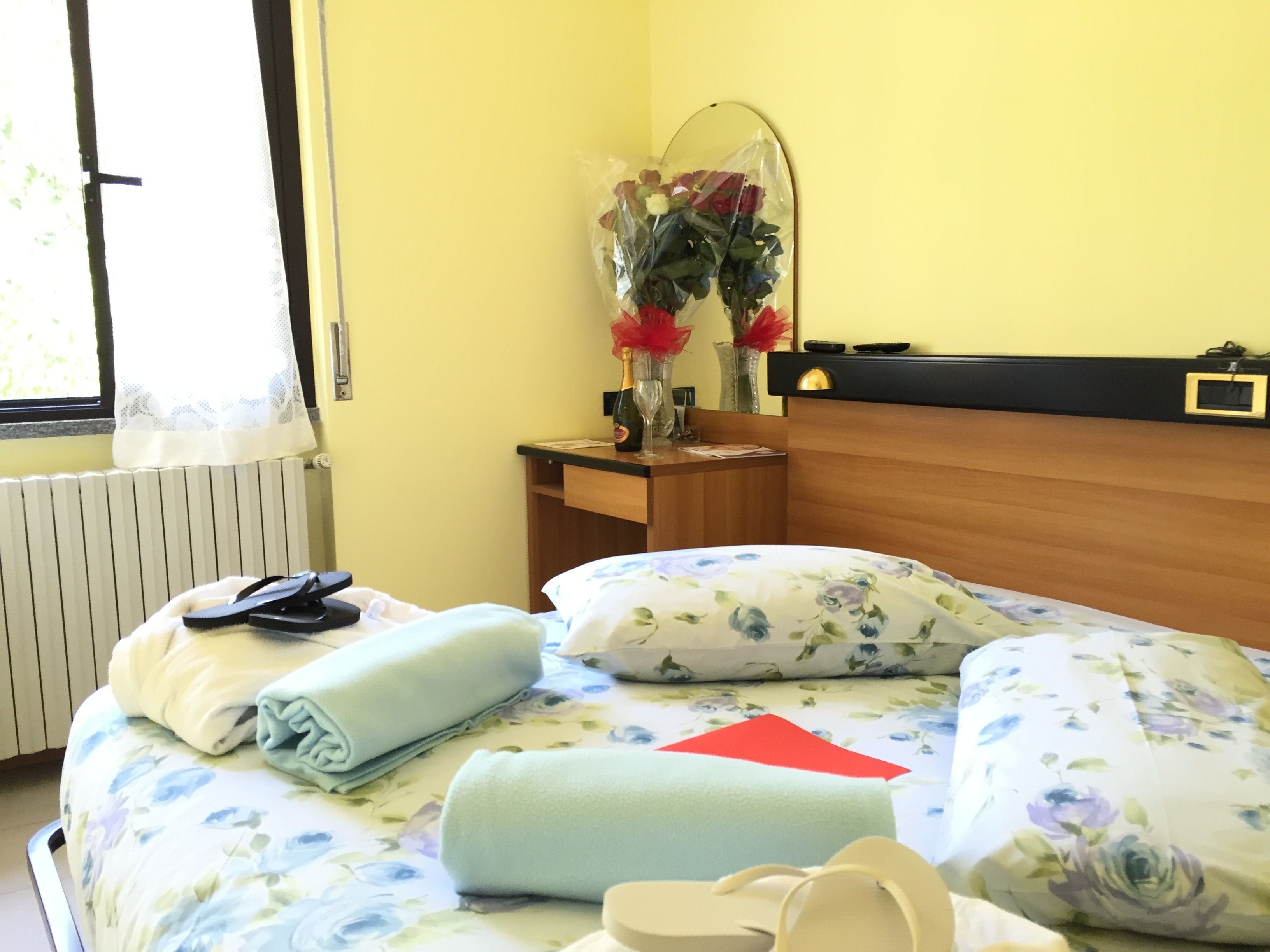 A hotel room with a bed, a nightstand, a mirror, and a window with a view of greenery.