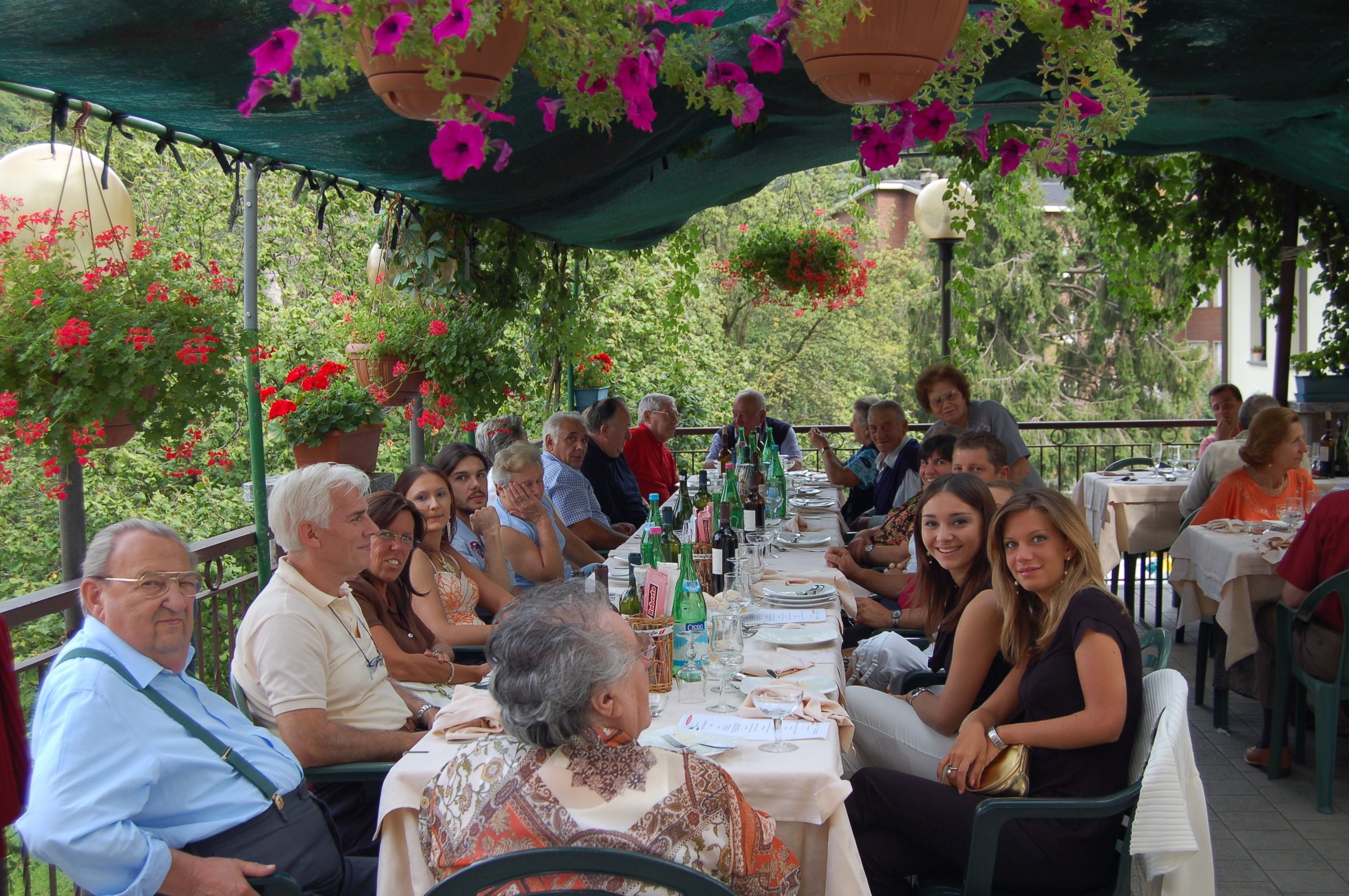 Un espacio de comedor al aire libre con mesas y sillas, rodeado de follaje y macetas colgantes de flores.