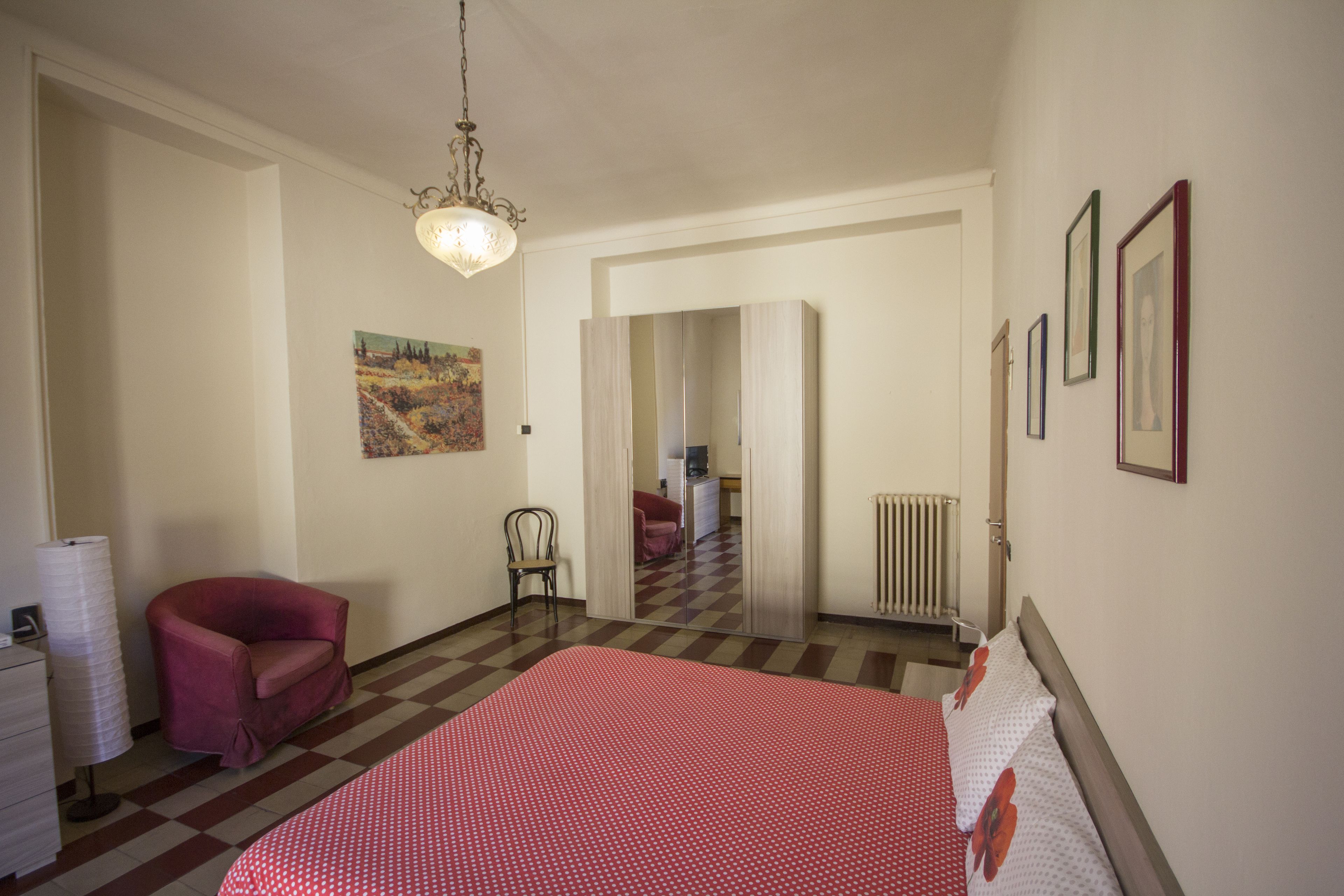 Ein Schlafzimmer mit kariertem Boden, einem Bett mit roten und weißen Punkten, einem roten Sessel, einem kleinen Schreibtisch mit Lampe und einem Bild an der Wand.