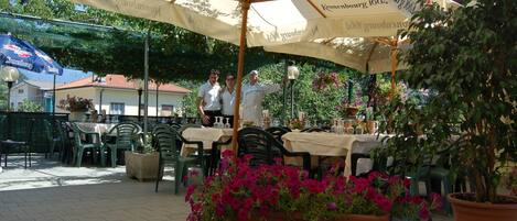 An outdoor café with tables and chairs, a large umbrella, and potted plants.