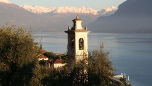 A tower with a bell, surrounded by trees and a lake with mountains in the background. - Albergo Sala (Valbrona)