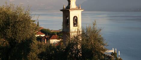 Una torre con una campana, rodeada de árboles y un lago con montañas al fondo.