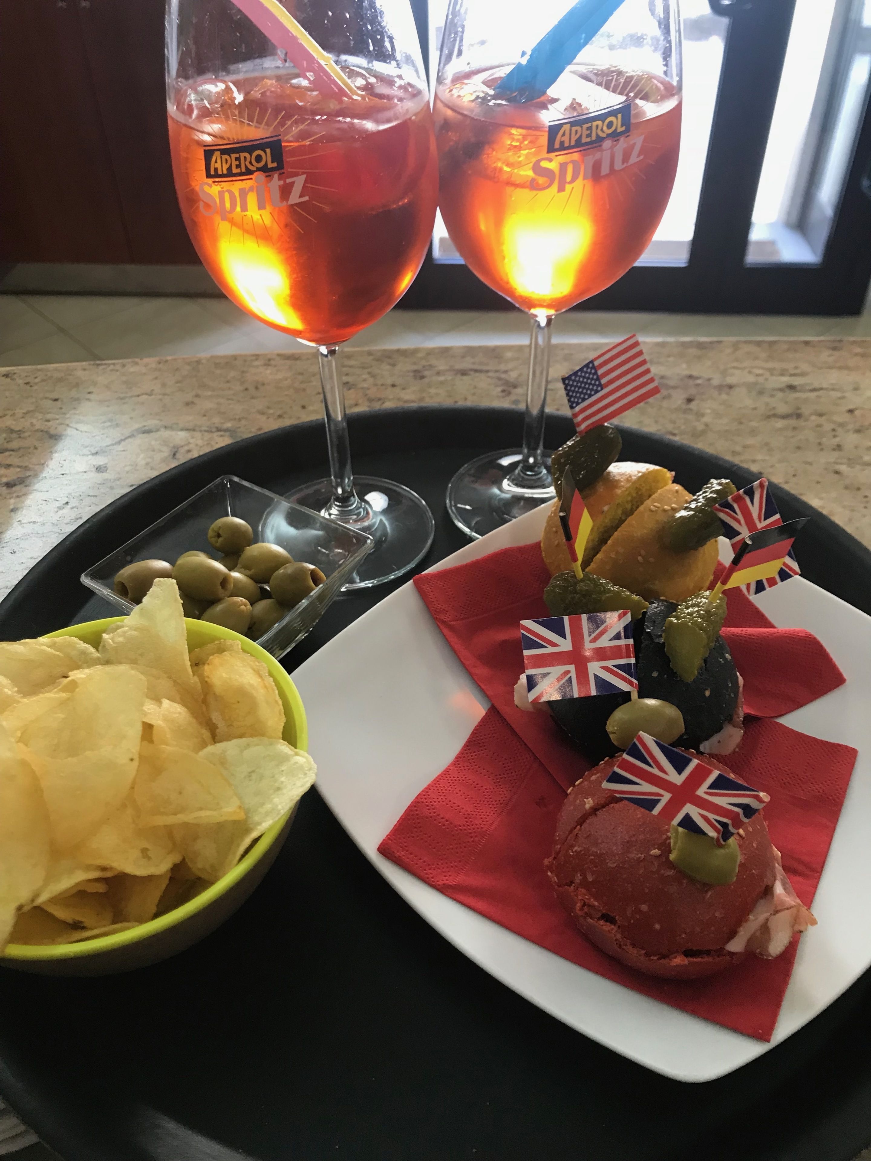 A tray with two glasses of Aperol Spritz, a plate with a sandwich and pickles, and a bowl of chips.