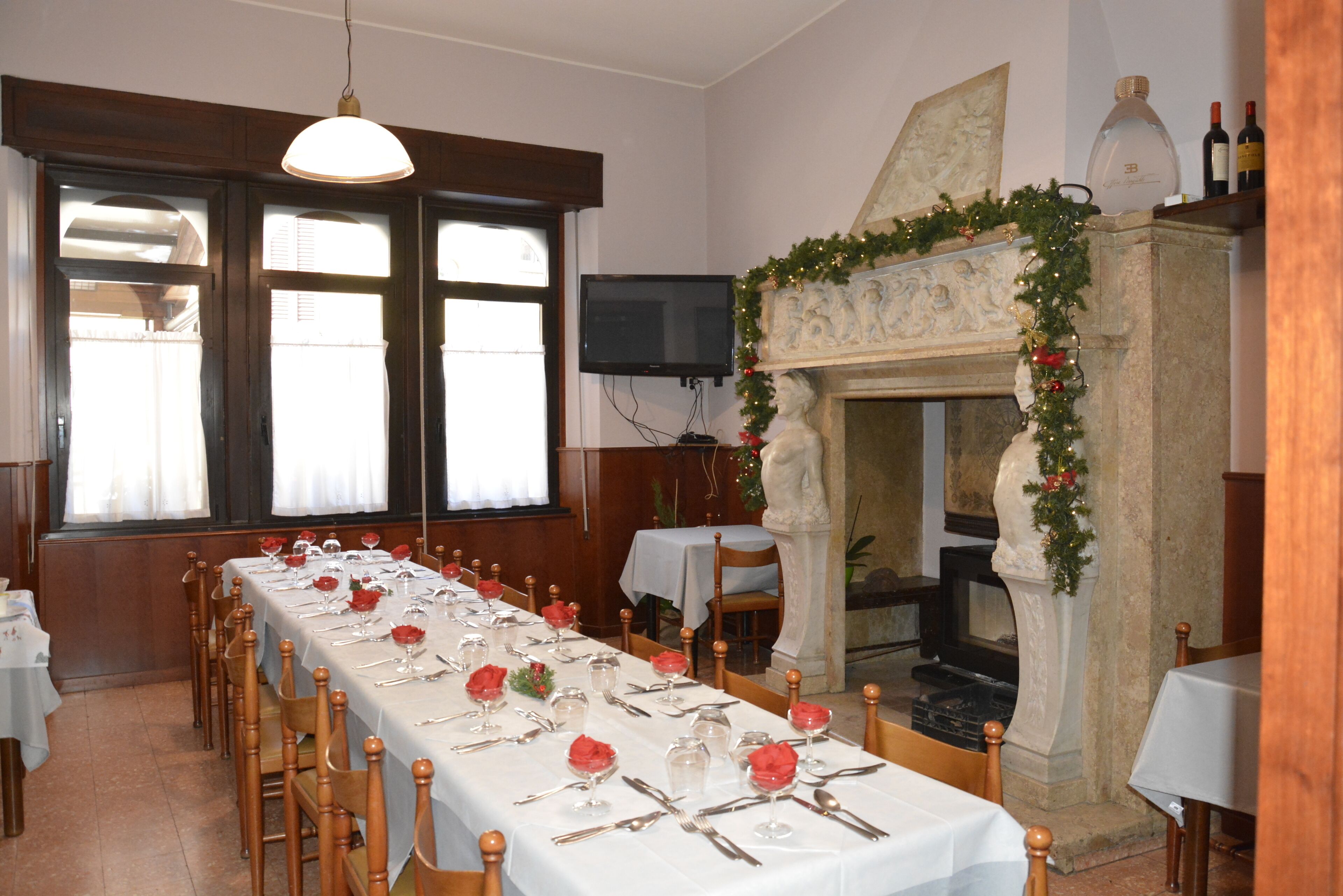 Ein langer Esstisch, gedeckt mit roten Blumen, Besteck und Weingläsern, mit Holzsesseln an den Seiten. Im Hintergrund ist ein Kamin mit Steinverkleidung und festlicher Girlande zu sehen.
