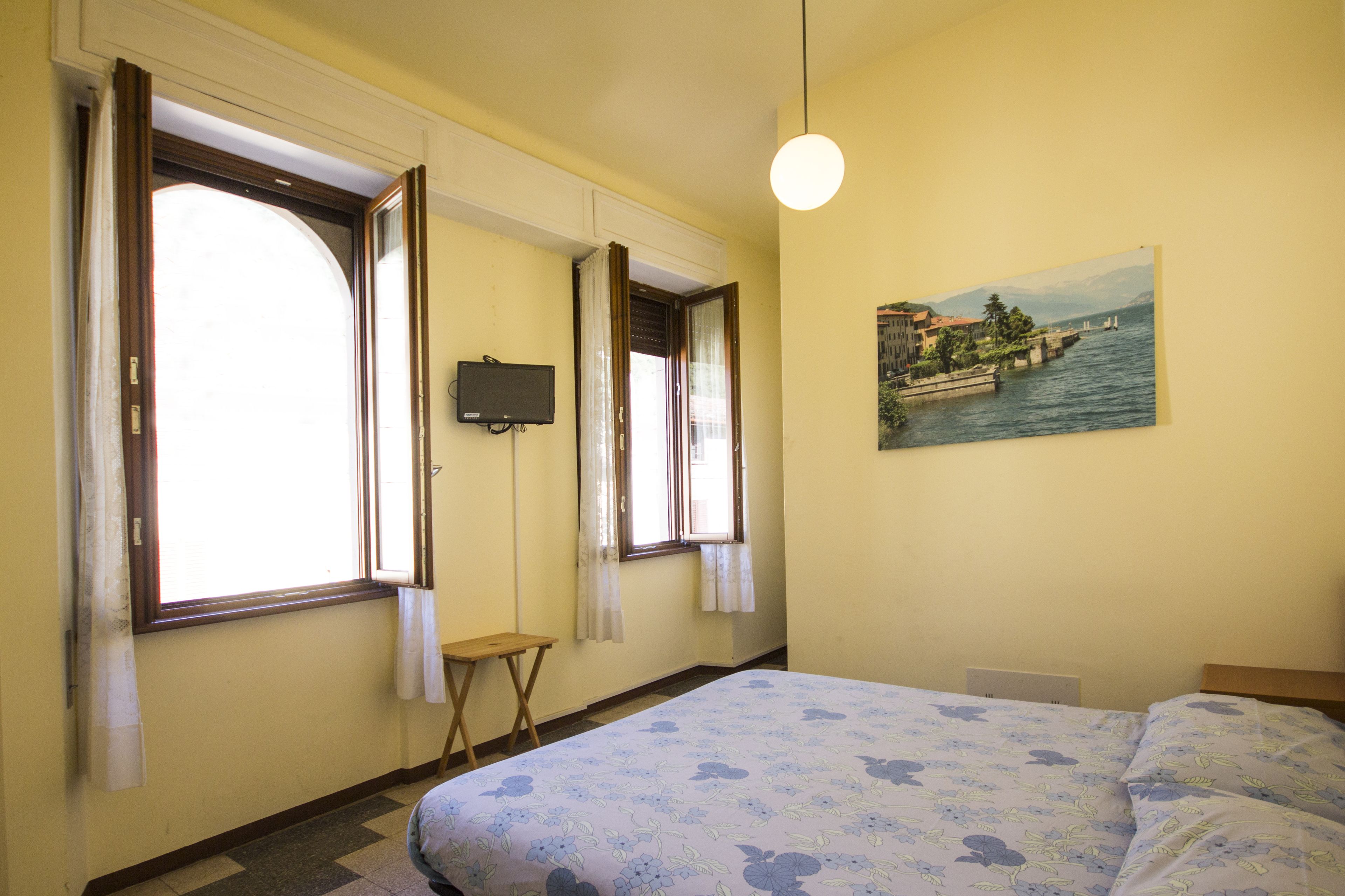Una habitación con una cama, dos ventanas con cortinas, una mesita pequeña, un televisor de pared y un cuadro de un paisaje lacustre.