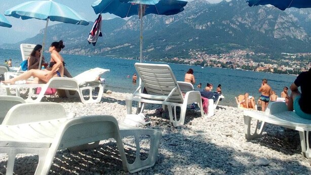 A beach with white plastic lounge chairs and blue umbrellas, a clear sky, and mountains in the background. - Albergo Sala (Valbrona)