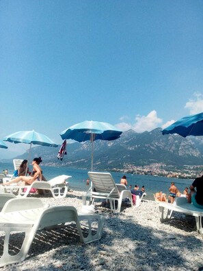 A beach with white plastic lounge chairs and blue umbrellas, a clear sky, and mountains in the background. - Albergo Sala (Valbrona)