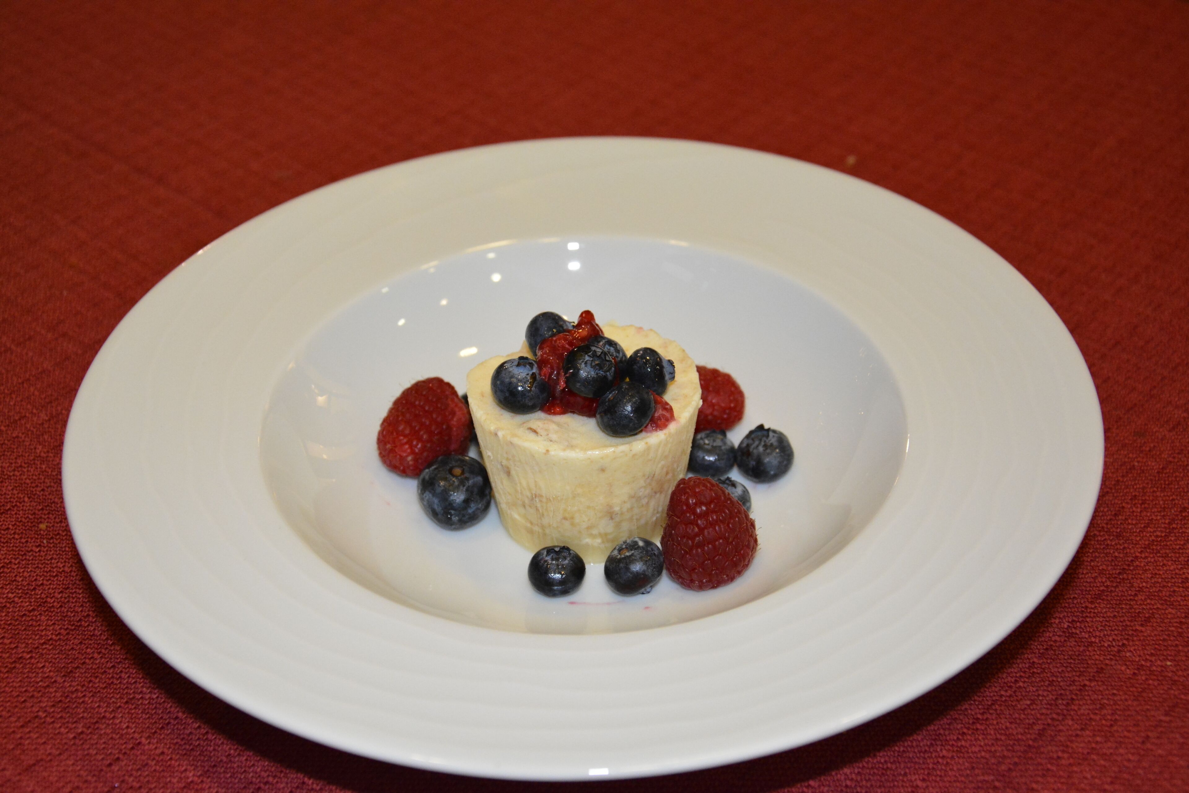 Un dessert servi sur une assiette blanche, agrémenté de myrtilles et de framboises, le tout posé sur une nappe rouge.