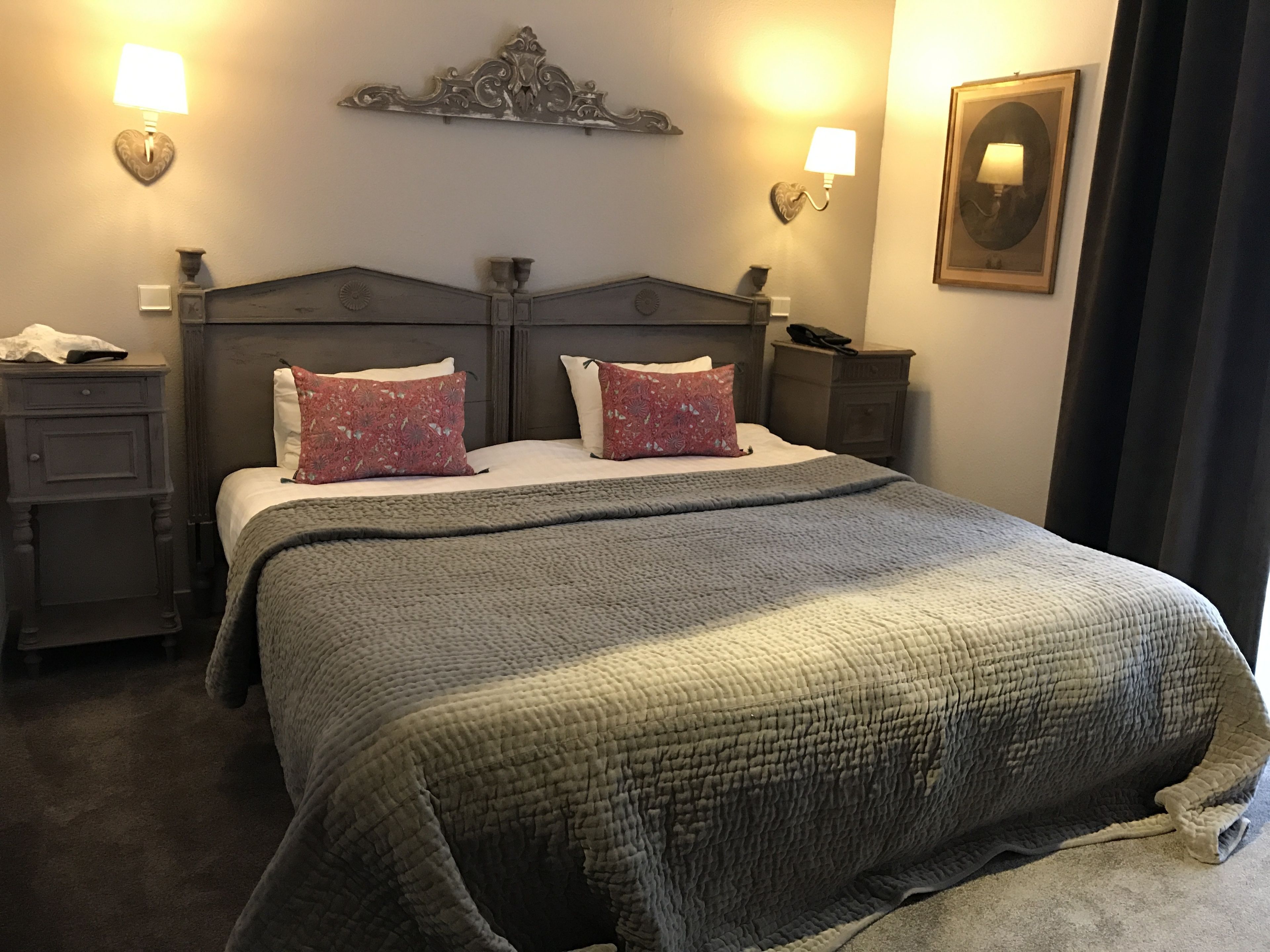 Een net opgemaakt bed met een grijze deken, twee rode kussens met een patroon en een decoratief hoofdbord. Er staan nachtkastjes met lampen en er hangt een ingelijste foto aan de muur.