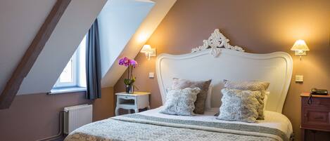 A bedroom with a sloped ceiling, a large bed with a decorative headboard, two bedside lamps, a nightstand with a vase of flowers, and a dresser.