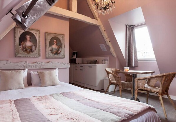 A bedroom with a large bed, a dining table, wicker chairs, a kitchenette, and two framed portraits on the wall.
