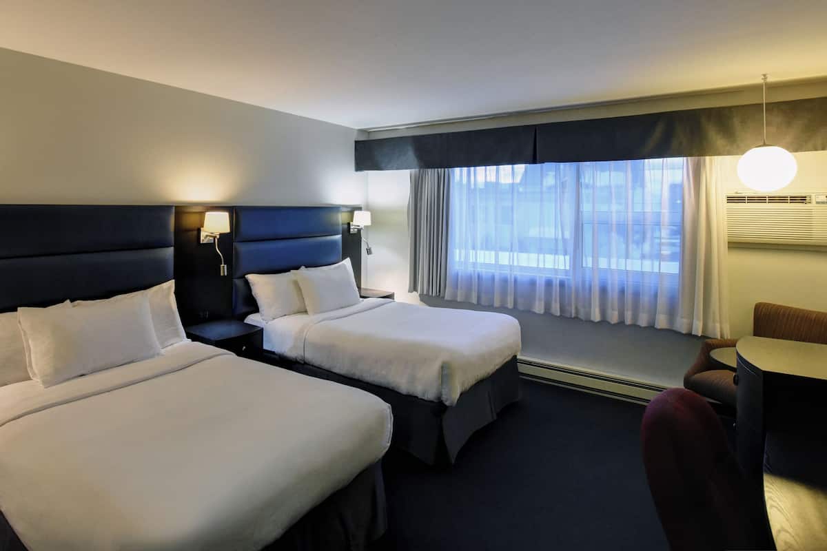 Una habitación de hotel con dos camas, un escritorio, una silla, una lámpara y un ventanal con cortinas.