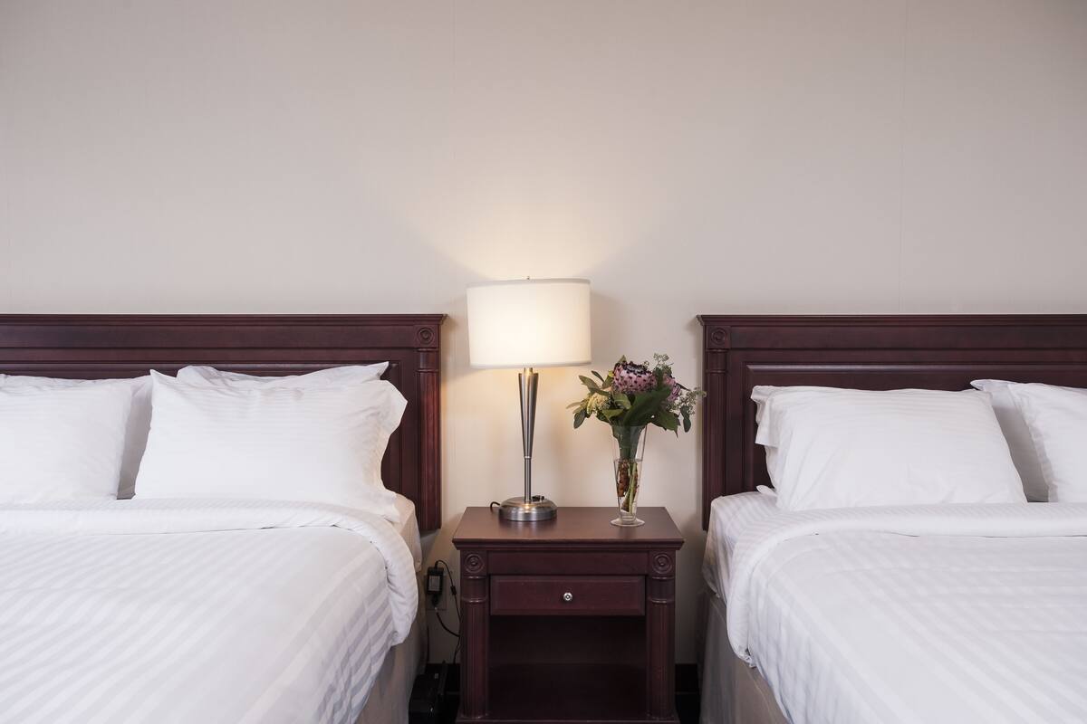 Una habitación de hotel con dos camas, una mesita de noche con lámpara y un jarrón con flores.
