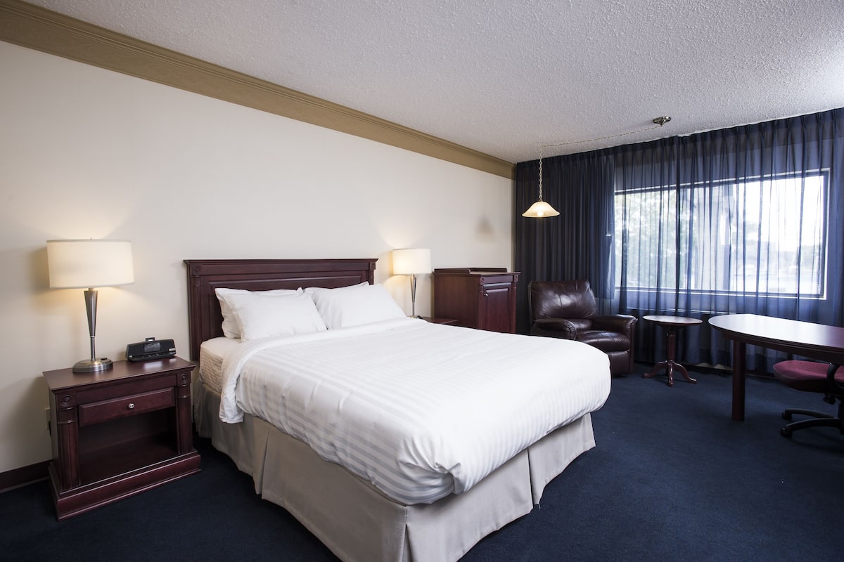 Una habitación de hotel con una cama grande, dos mesitas de noche con lámparas, un sillón de cuero, una pequeña mesa redonda y un gran ventanal con cortinas.