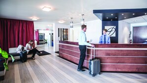 Zona de recepción de hotel con un mostrador de madera, un área de espera con un sofá y una silla, y una persona de pie con una maleta.