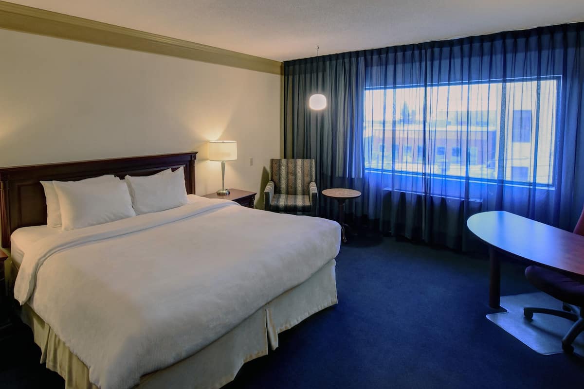 Habitación de hotel con una cama grande, dos lámparas de noche, una silla, una mesa y vistas de la ciudad a través de la ventana.