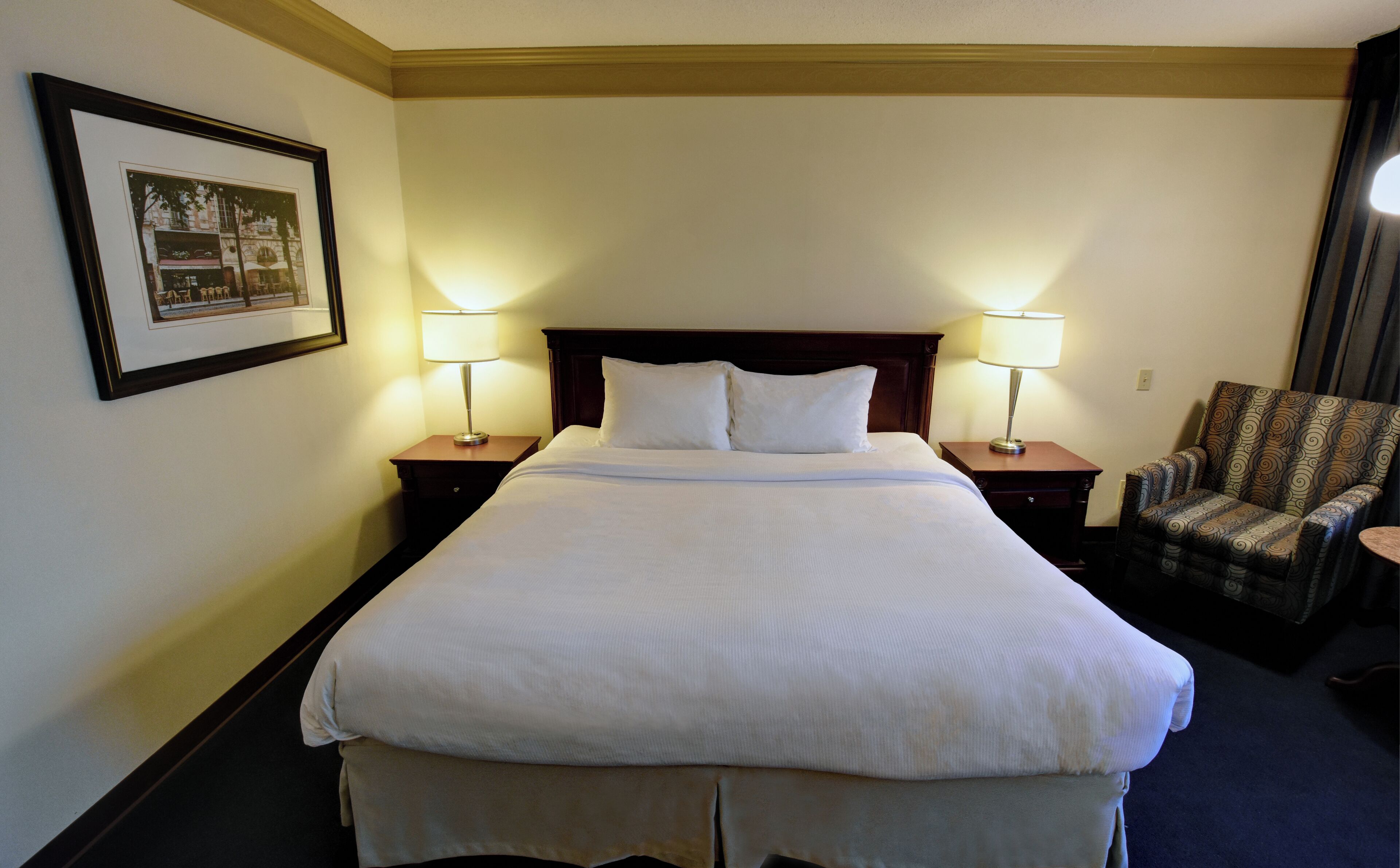 A neatly made bed with white linens, two bedside lamps, a framed picture on the wall, and a patterned armchair.