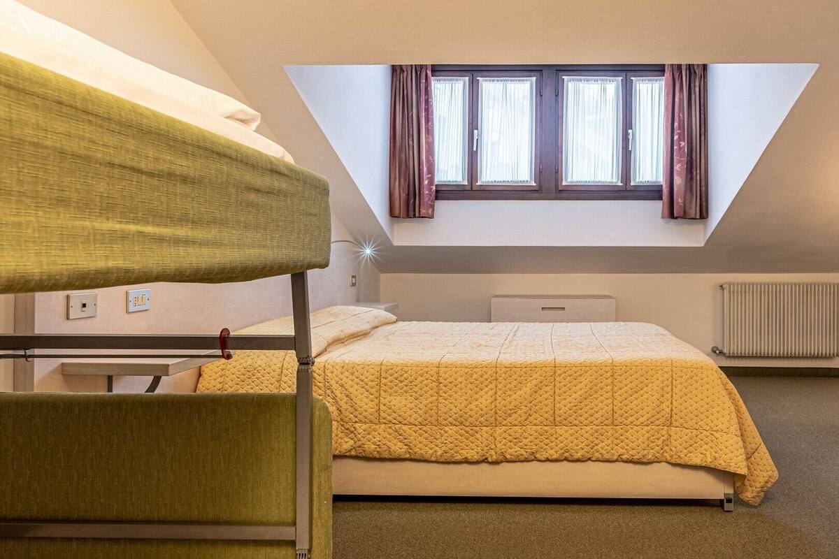 A dormitory room with bunk beds, a single bed, a skylight, a radiator, and a window with curtains.