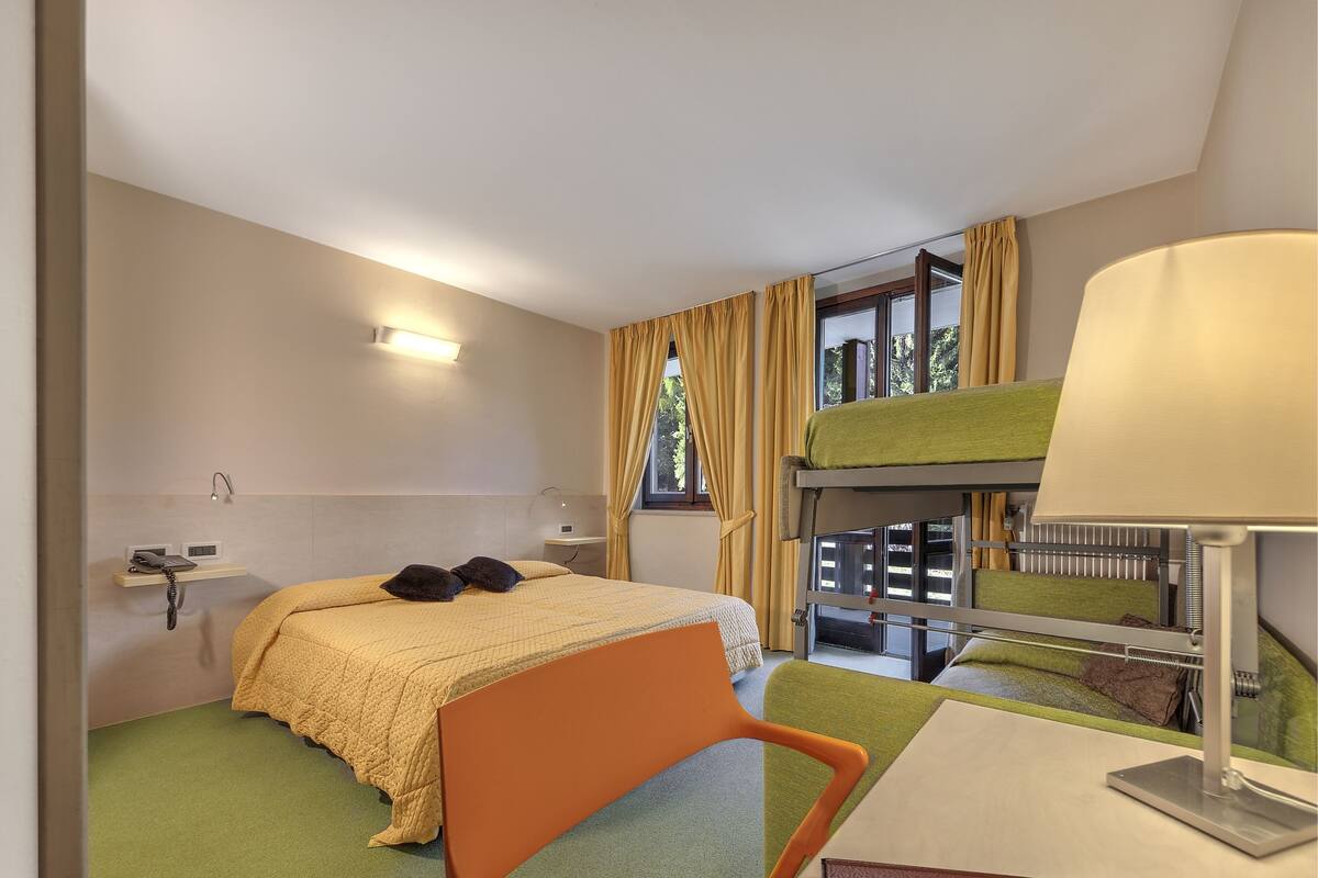 A compact hotel room with a bunk bed, a desk with a lamp, a chair, a bedside table with a phone, and a window with curtains.