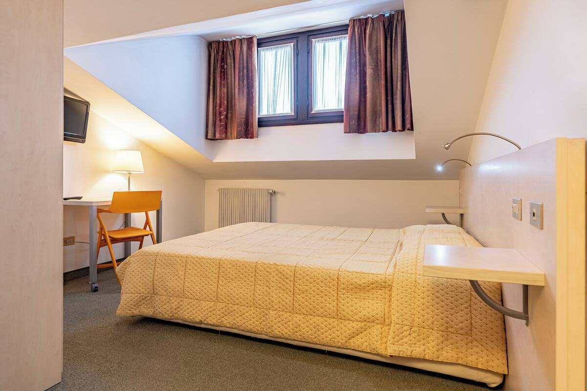A small, well-lit room with a bed, a desk, and a chair. There is a window with curtains, a radiator, and a TV mounted on the wall.