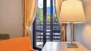 Habitación de hotel con un ventanal grande, cortinas, una mesa con lámpara, una silla y un control remoto.