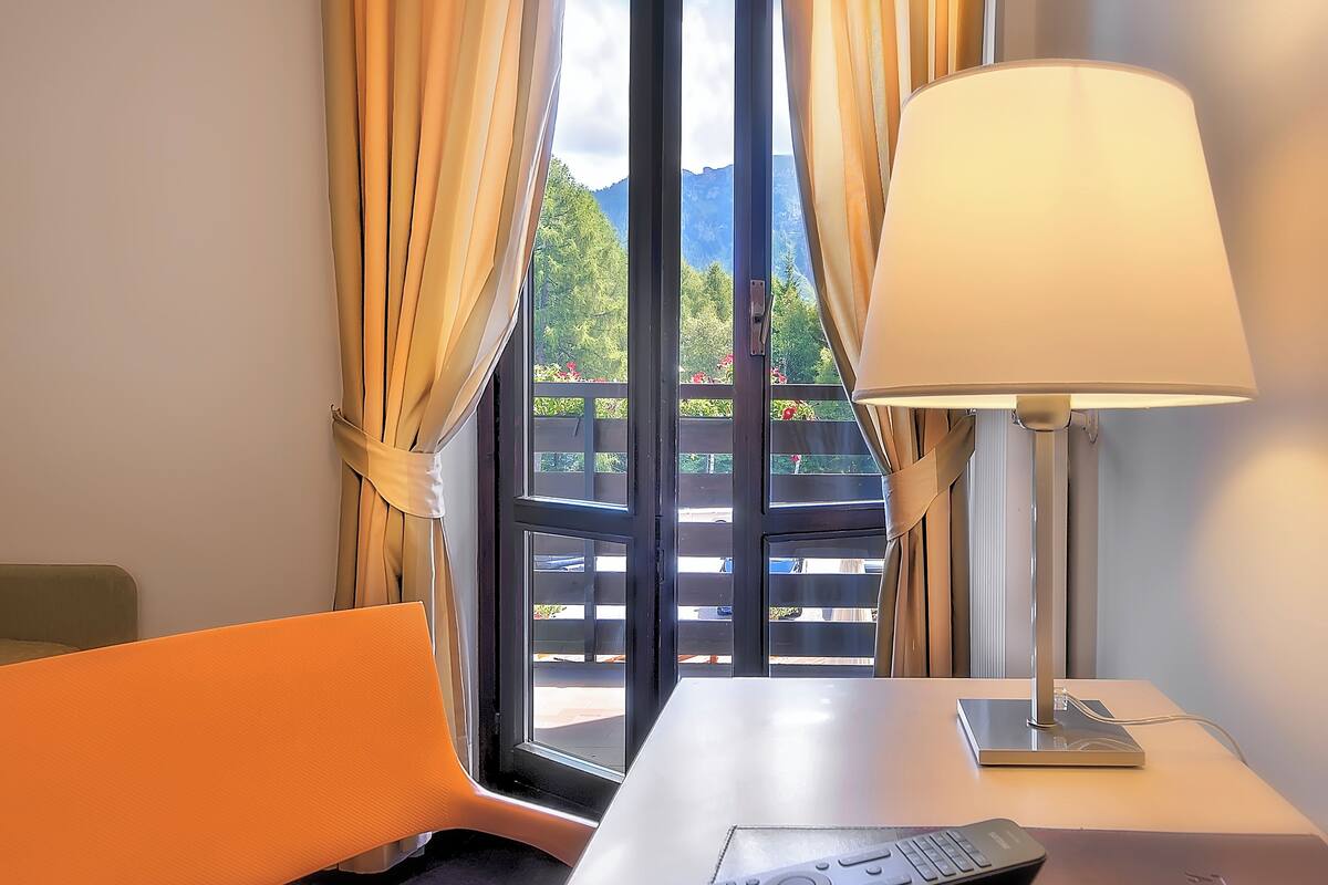 A hotel room with a large window, curtains, a table with a lamp, a chair, and a remote control.