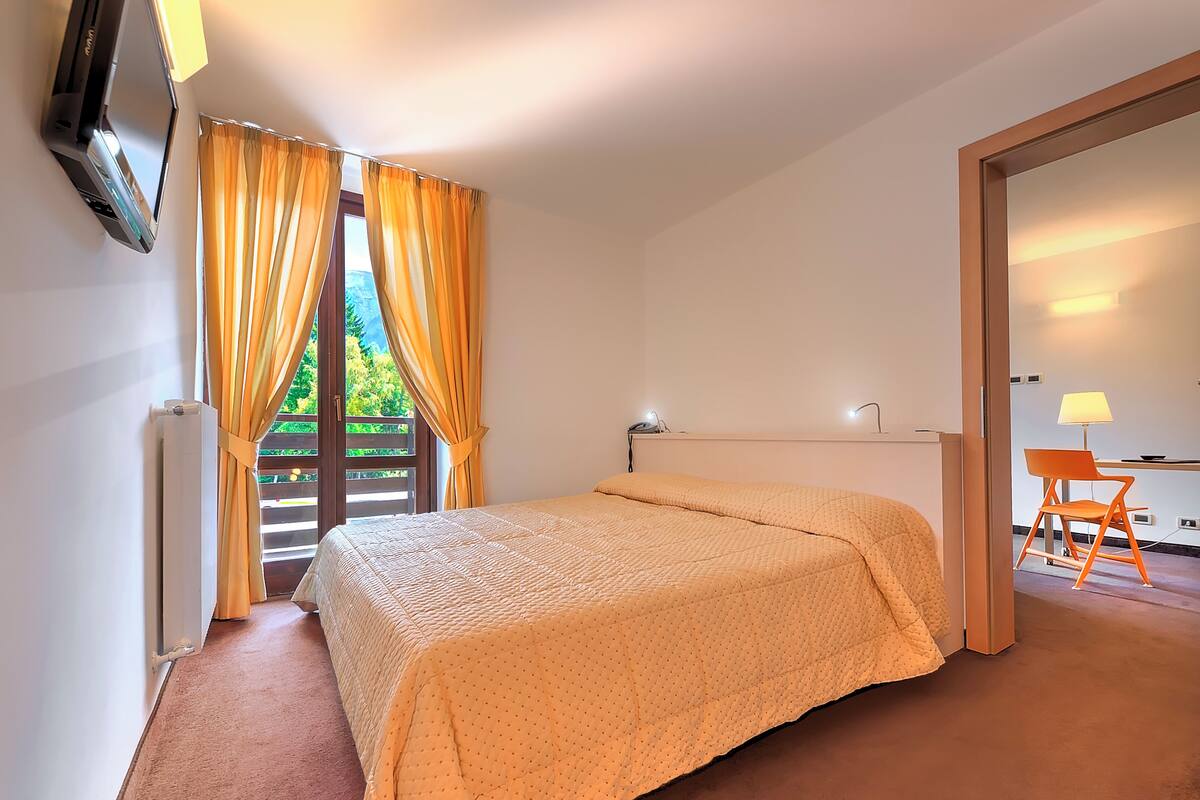 A bedroom with a bed, a desk with a chair, a radiator, and curtains.