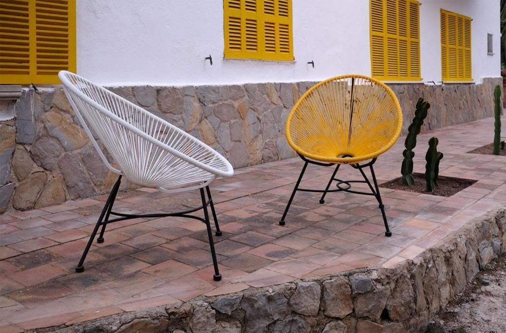Two chairs with metal frames and woven seats, placed on a stone-paved patio with a white wall and yellow shutters in the background.