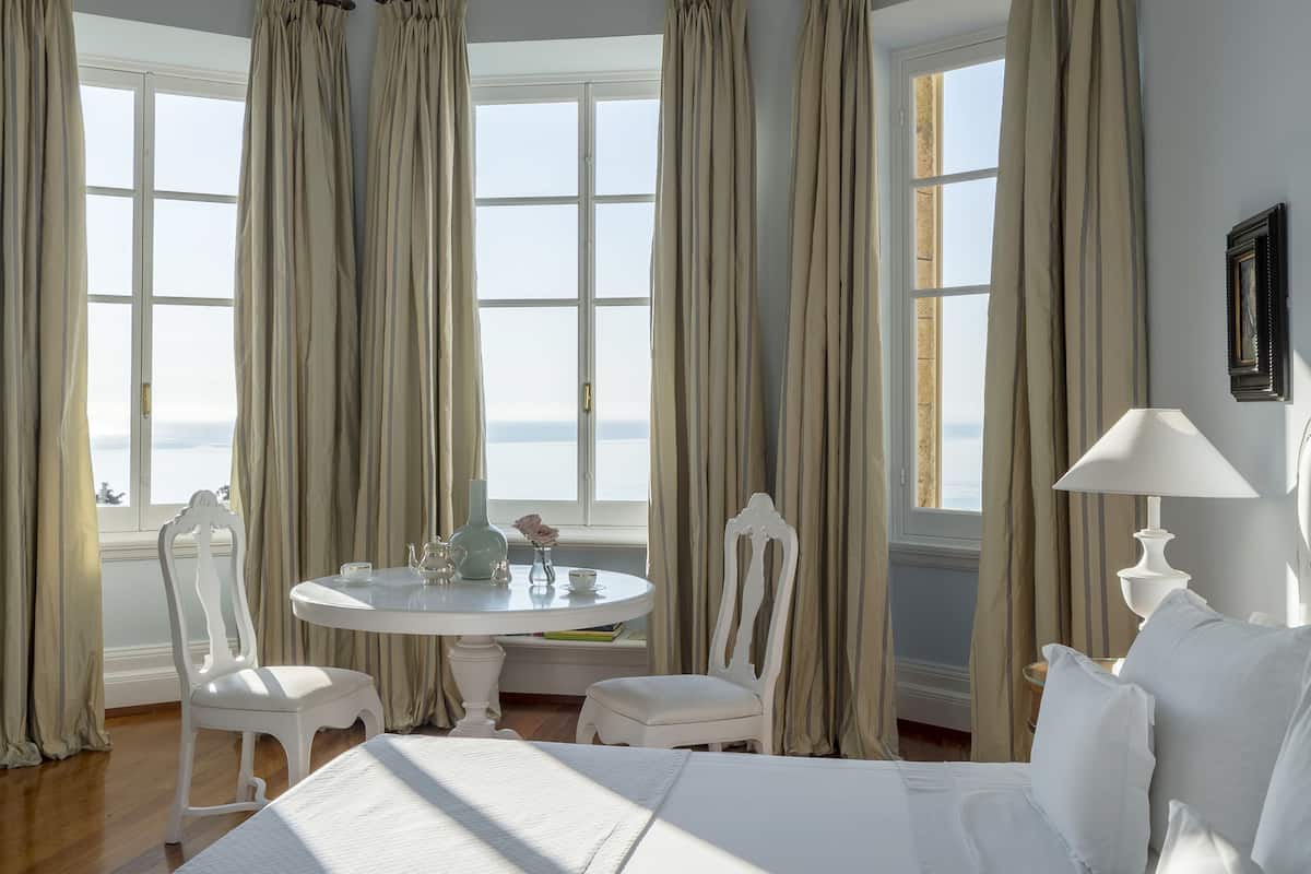 A hotel room with a large bed, a dining table with chairs, and a view of the sea through large windows.