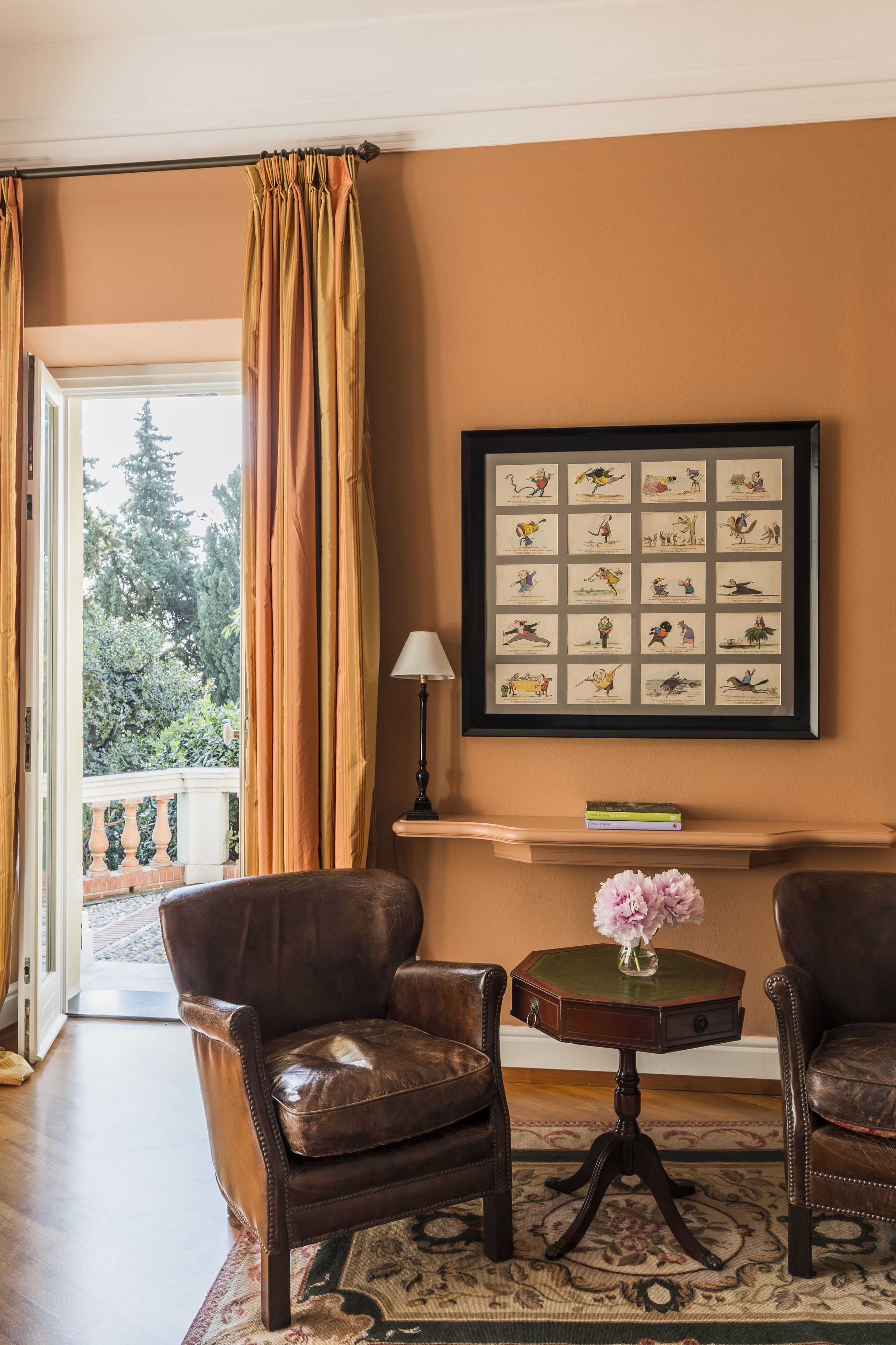 Una habitación con un ventanal grande, un cuadro enmarcados con ilustraciones, dos sillones de cuero, una mesita con un florero y una lámpara.