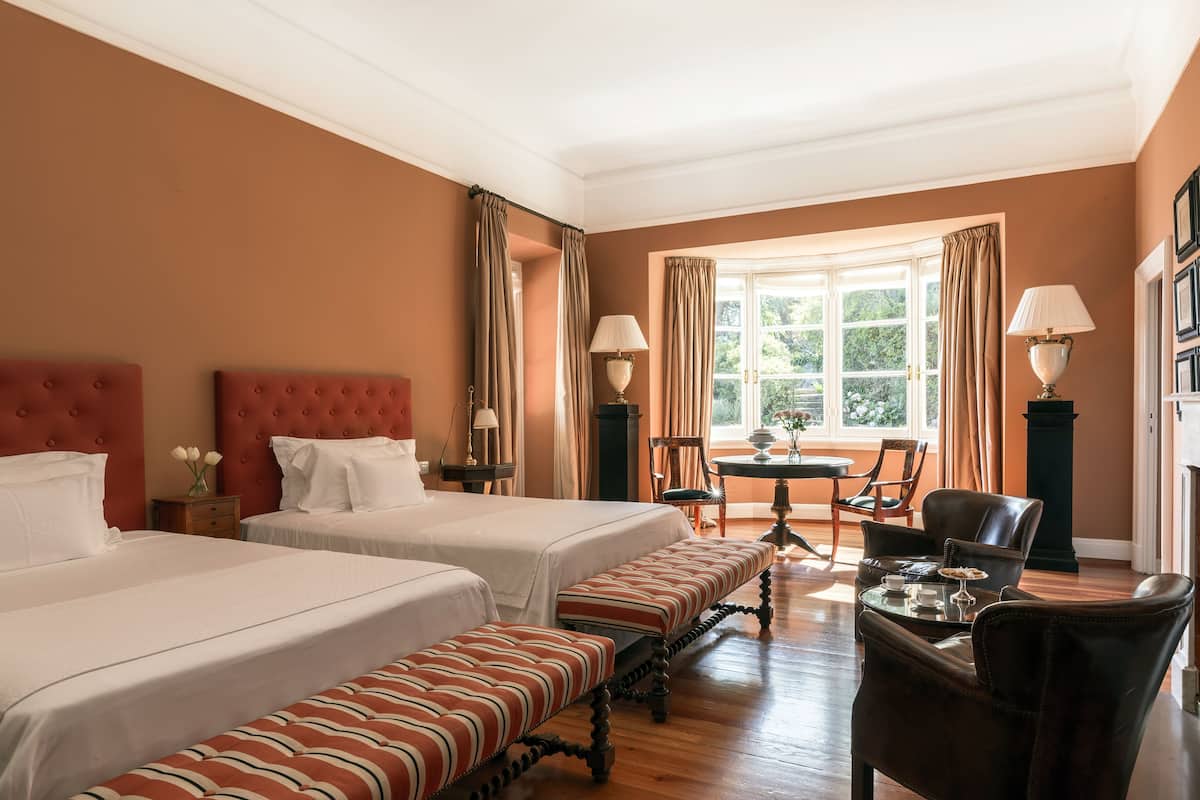 A hotel room with two beds, a bench, a table with chairs, and a window with curtains.