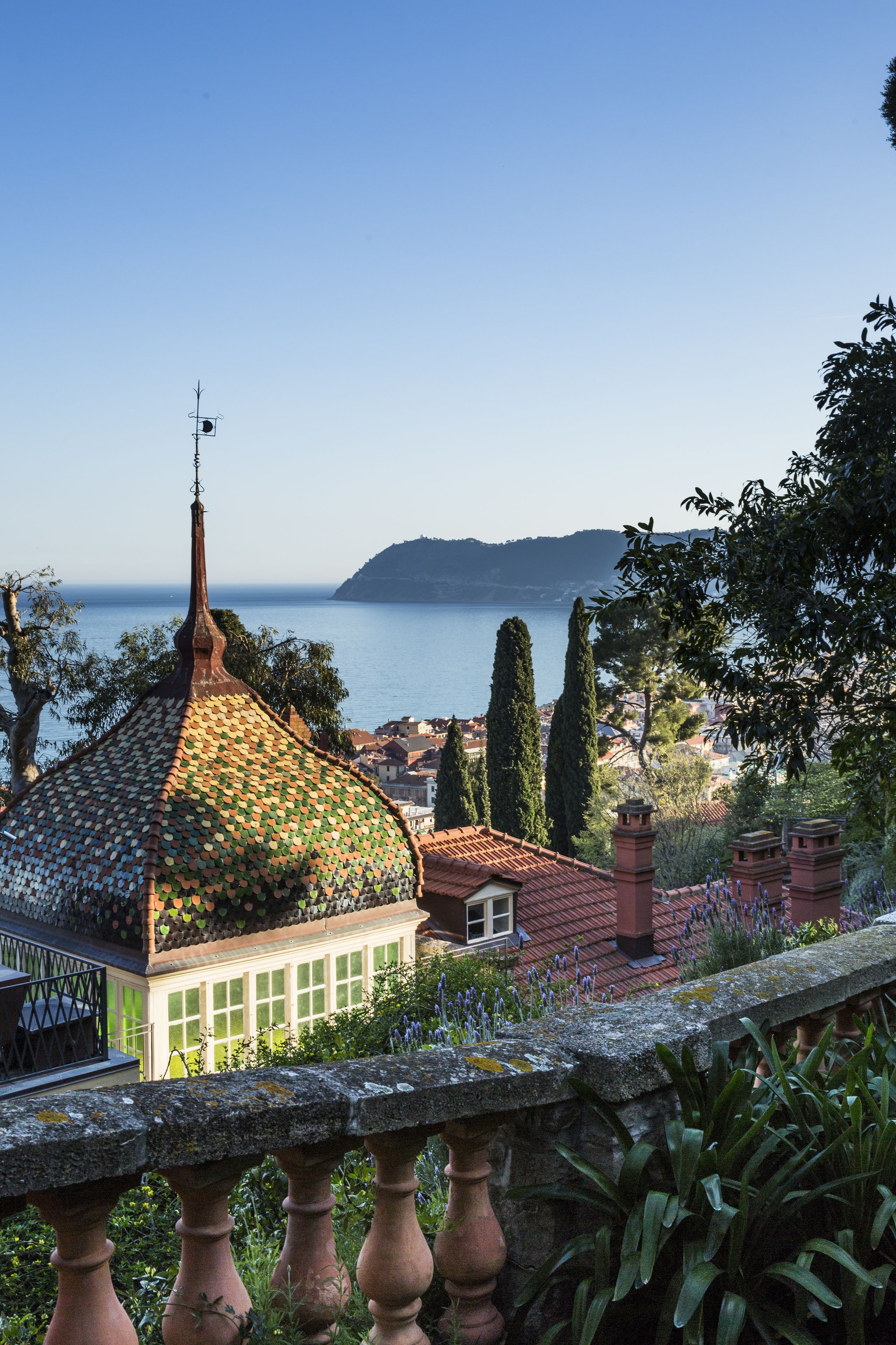 A building with a tiled dome roof, a stone balustrade, and a view of the sea and hills in the background.