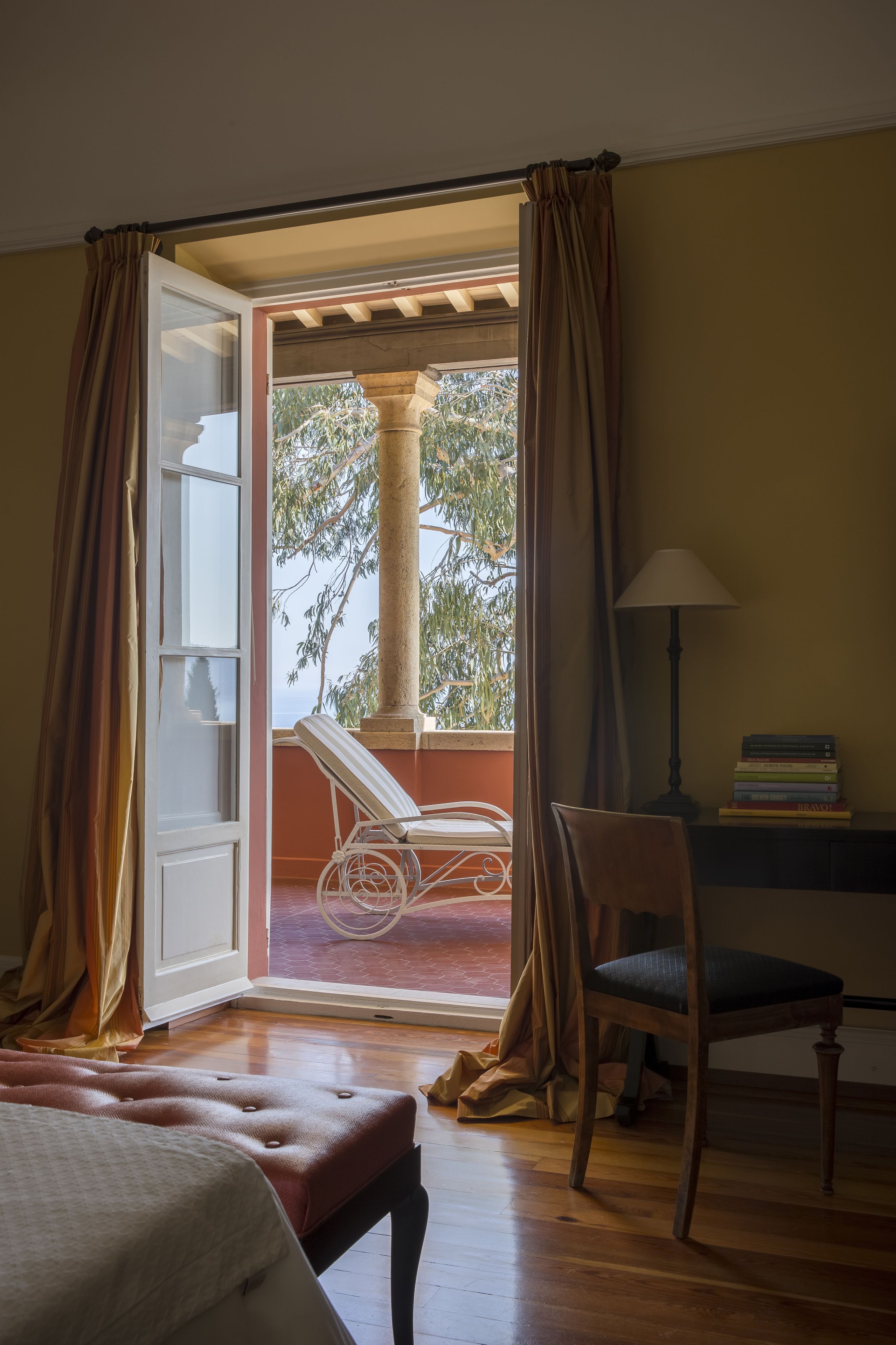 A room with a bed, a desk with a lamp, a chair, and an open door leading to a balcony with a chair and a table.