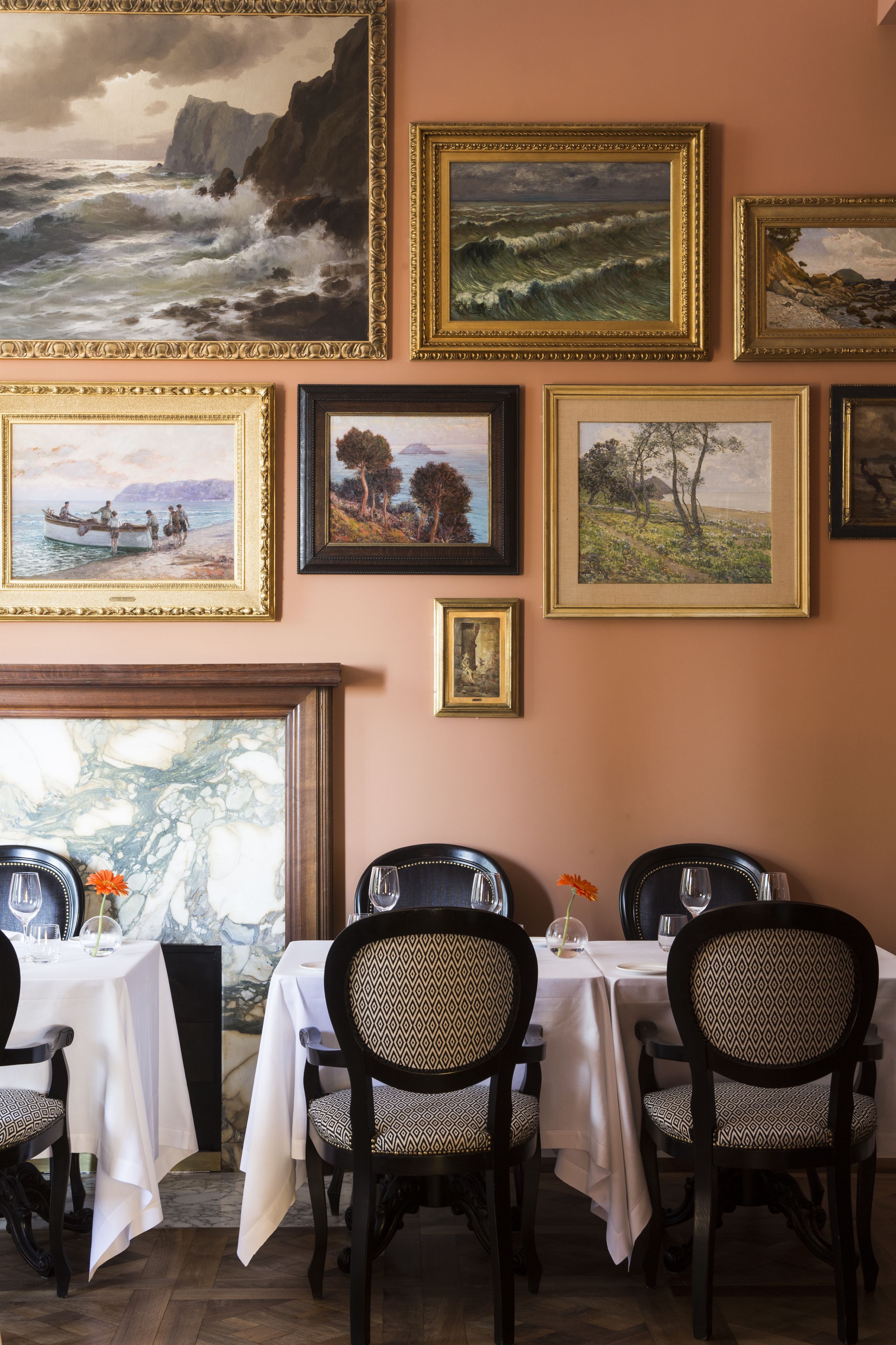 Un coin repas avec des tables disposées pour quatre personnes, des œuvres d’art encadrées accrochées au mur et une vue sur la mer par une fenêtre.