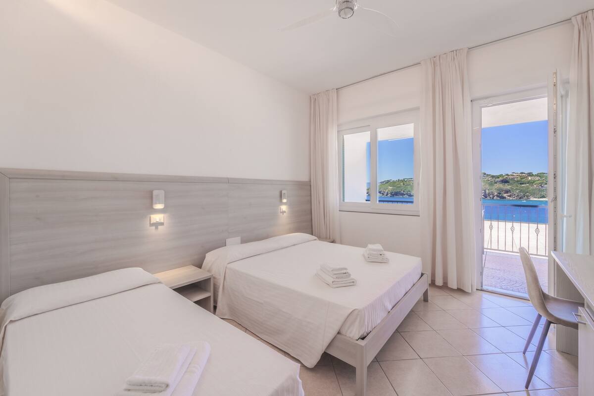 A hotel room with two beds, a desk, and a view of the sea.