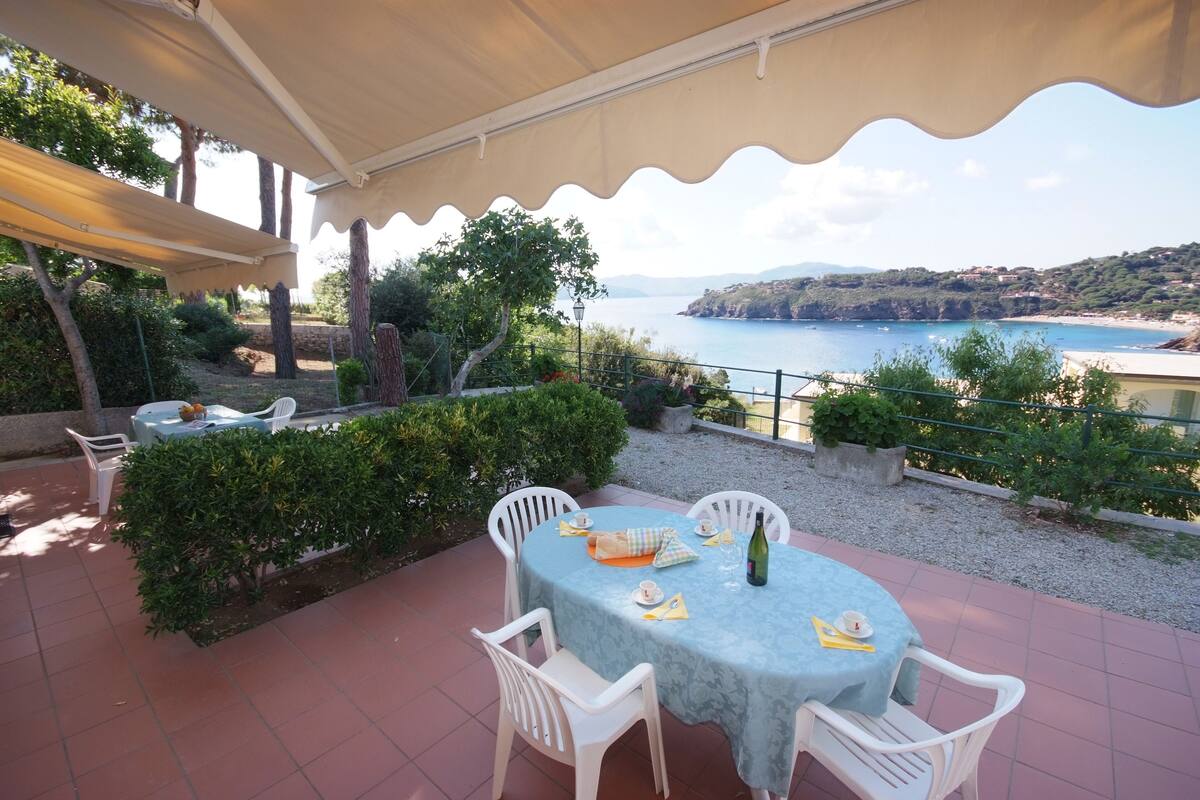 An outdoor patio with a table set for two, surrounded by white chairs, overlooking a coastal landscape.