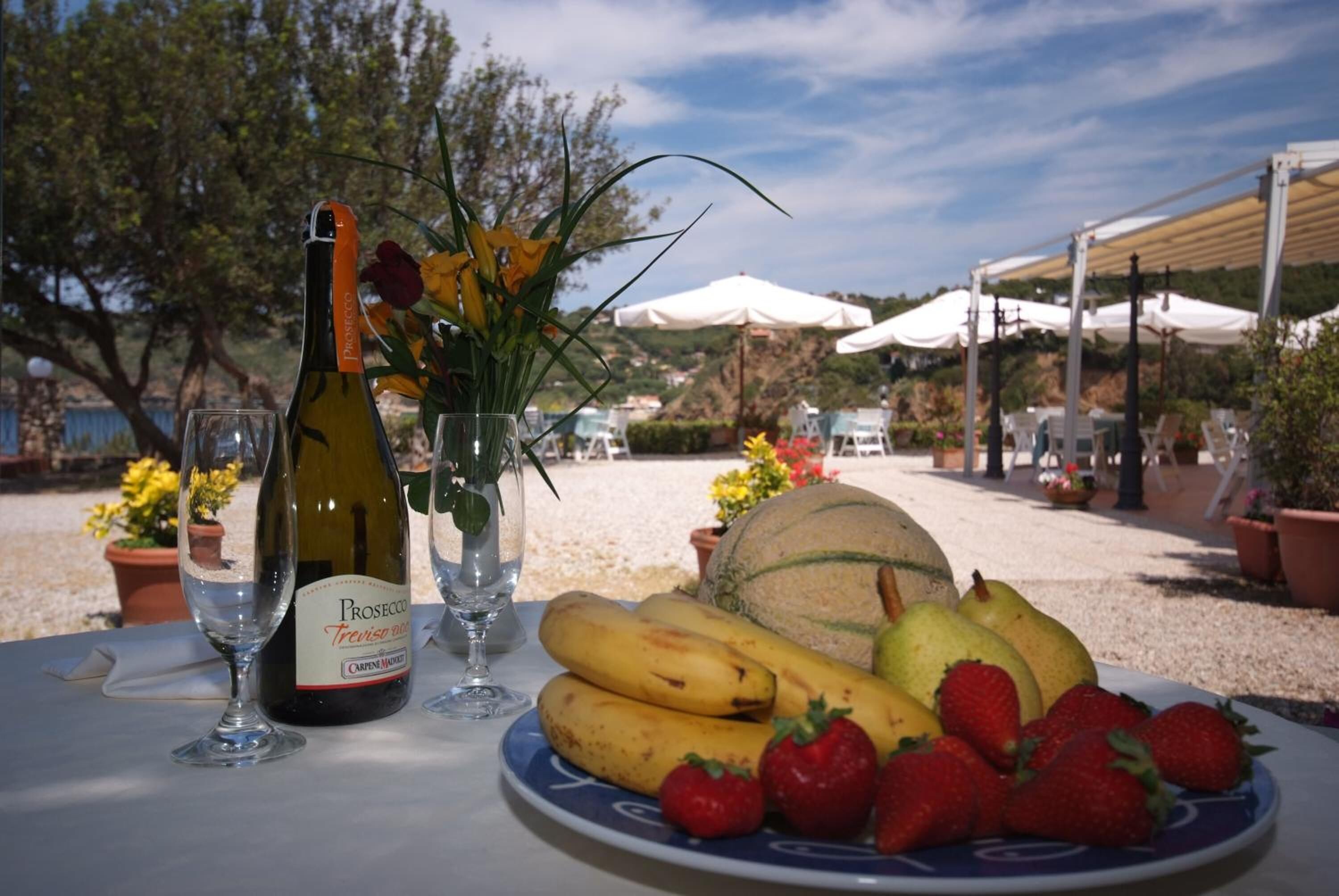 Una mesa con una botella de Prosecco, una copa, un florero con flores y un plato de fruta.