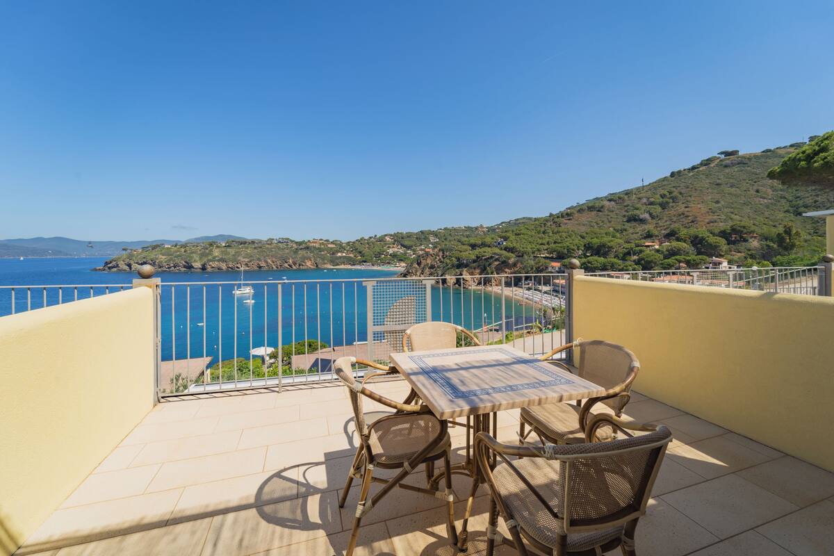 A balcony with wicker chairs and a table, overlooking a coastal area with a clear view of the sea and a hilly landscape.