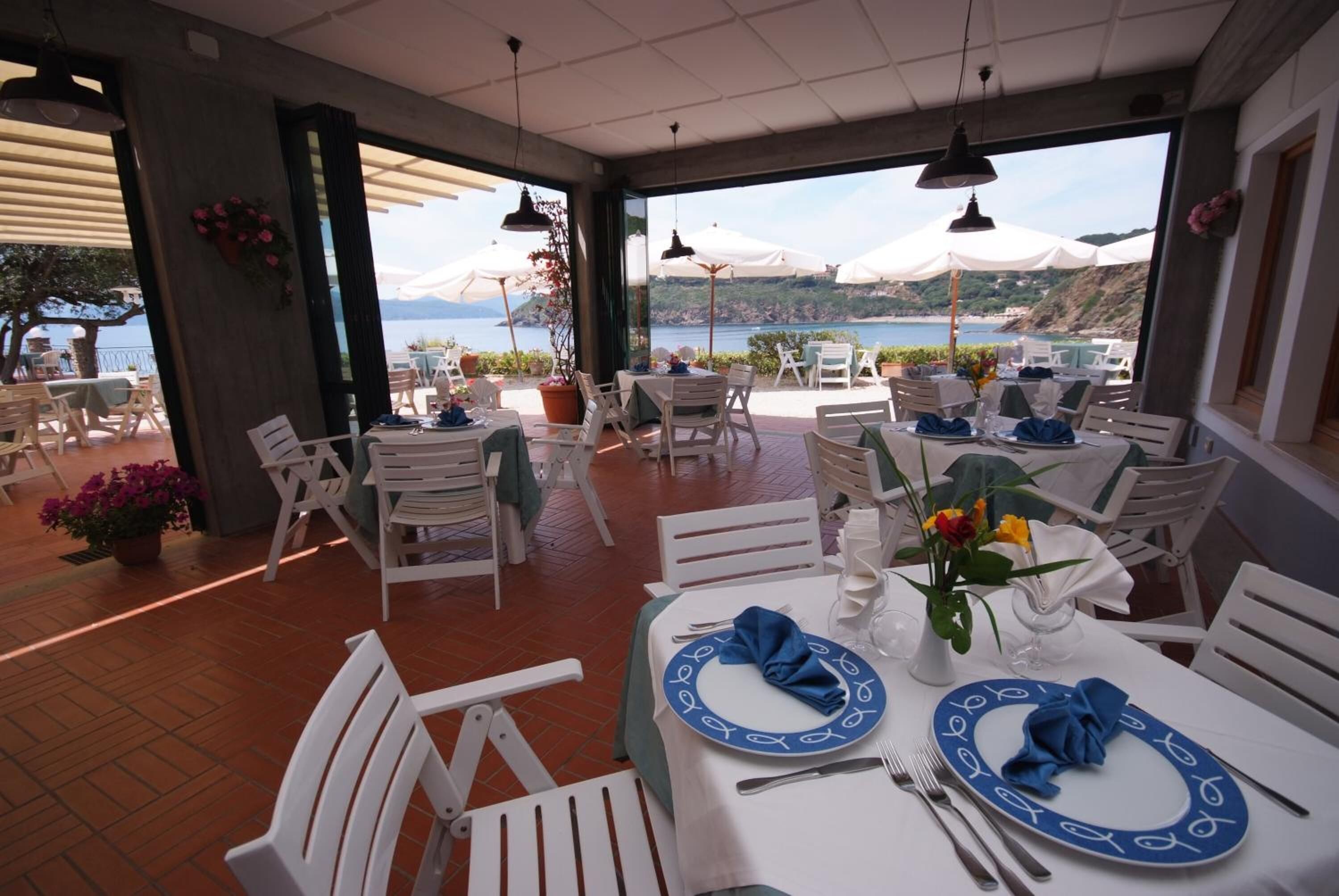 A restaurant with outdoor seating overlooking a scenic view of the sea and hills. Tables are set with blue and white tableware, and there are potted plants and umbrellas providing shade.