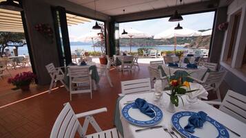 A restaurant with outdoor seating overlooking a scenic view of the sea and hills. Tables are set with blue and white tableware, and there are potted plants and umbrellas providing shade.
