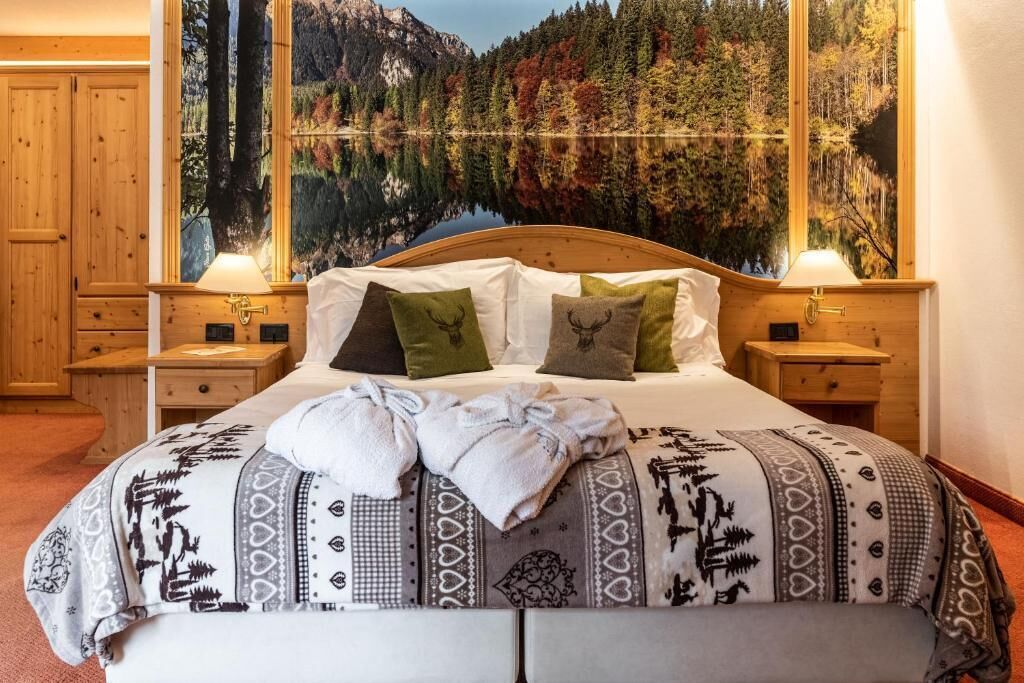 Una cama con una colcha estampada, dos almohadas, un par de toallas y un cabecero con una pintura de un bosque y un lago.