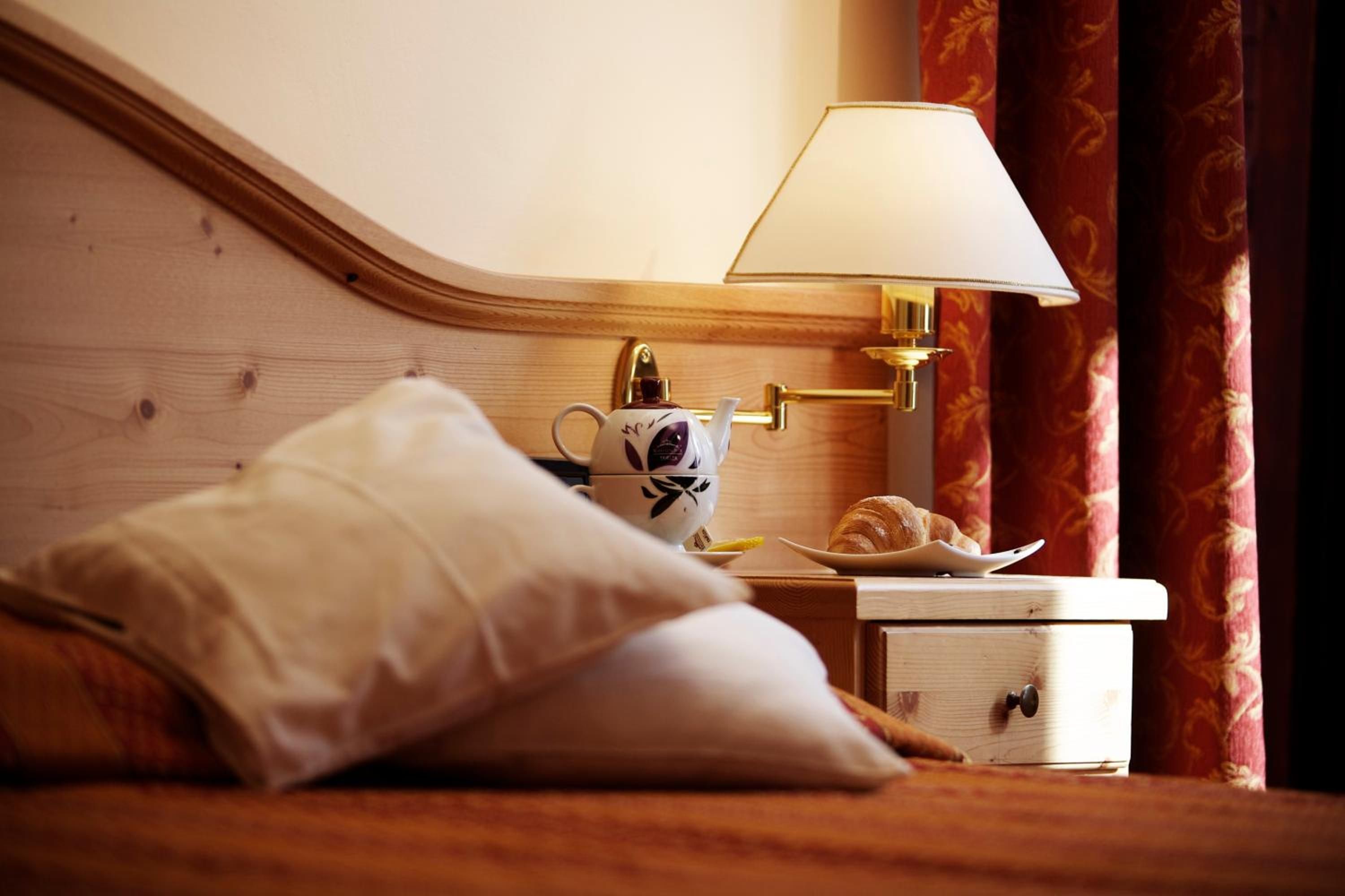 A neatly made bed with a pillow, a bedside table with a lamp, a teapot, and a croissant.