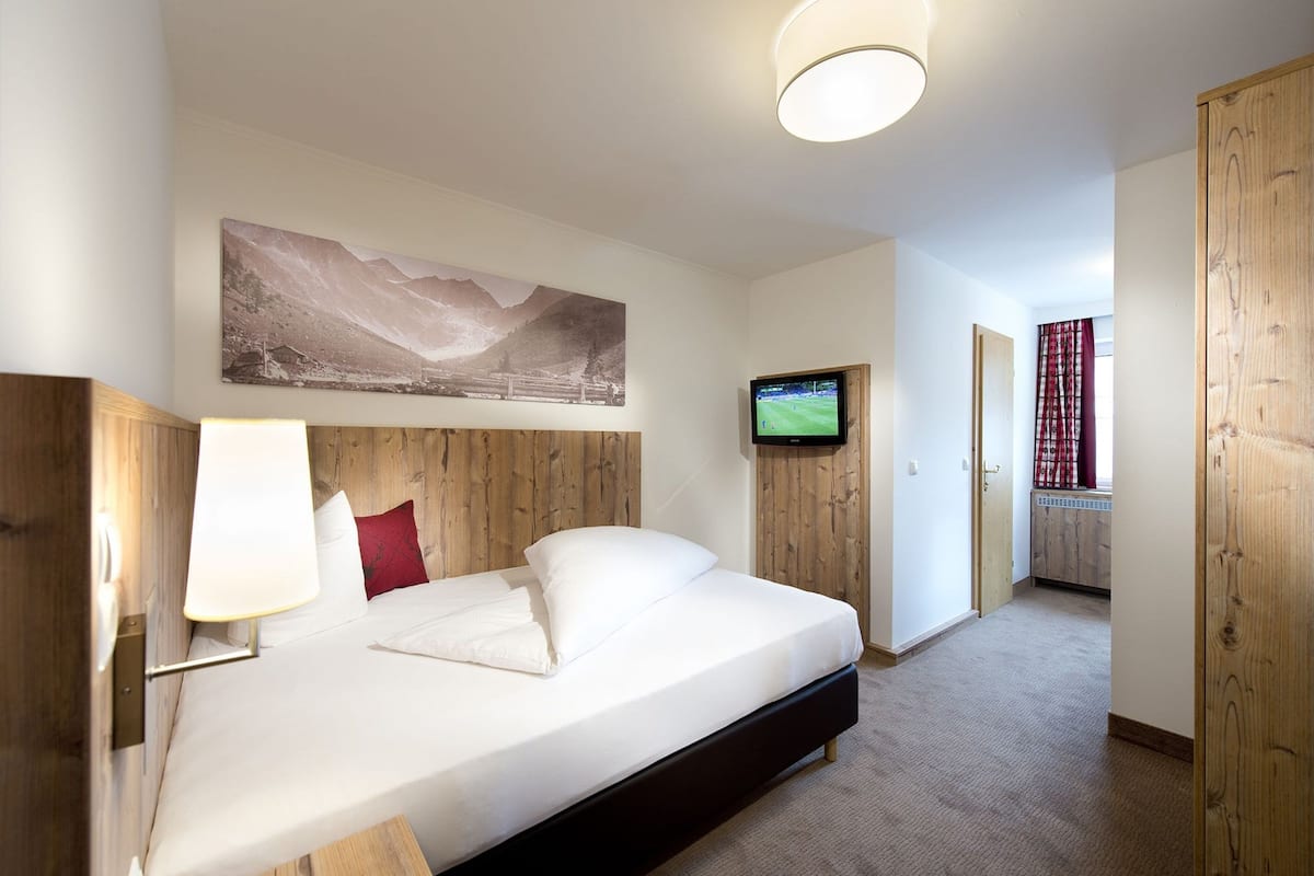 A bedroom with a bed, a mounted TV, a painting on the wall, and a wooden door.
