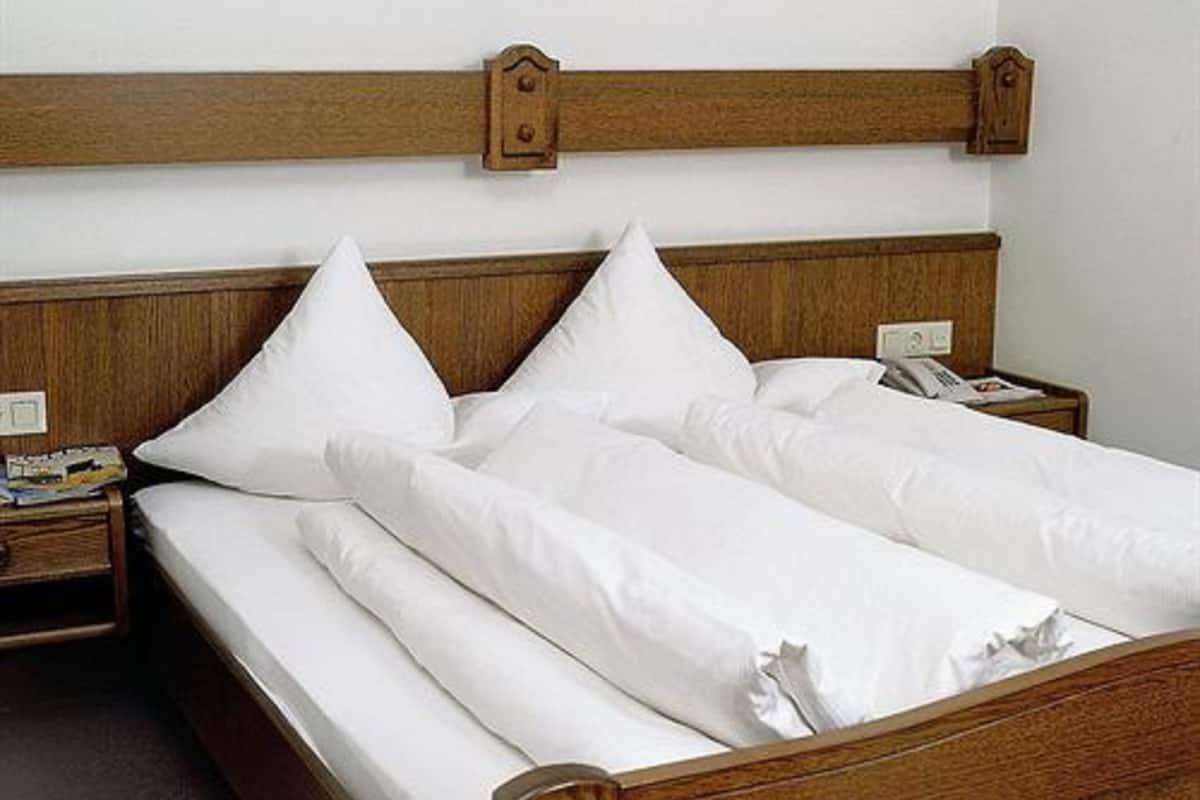 A neatly made bed with white linens and wooden headboard, a bedside table with a lamp, and a wall socket.