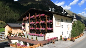 A mountain lodge with a curved roof, wooden balconies with flower boxes, and a sign reading "Dorfplatz". - Hotel Bergland (Sankt Leonhard im Pitztal)