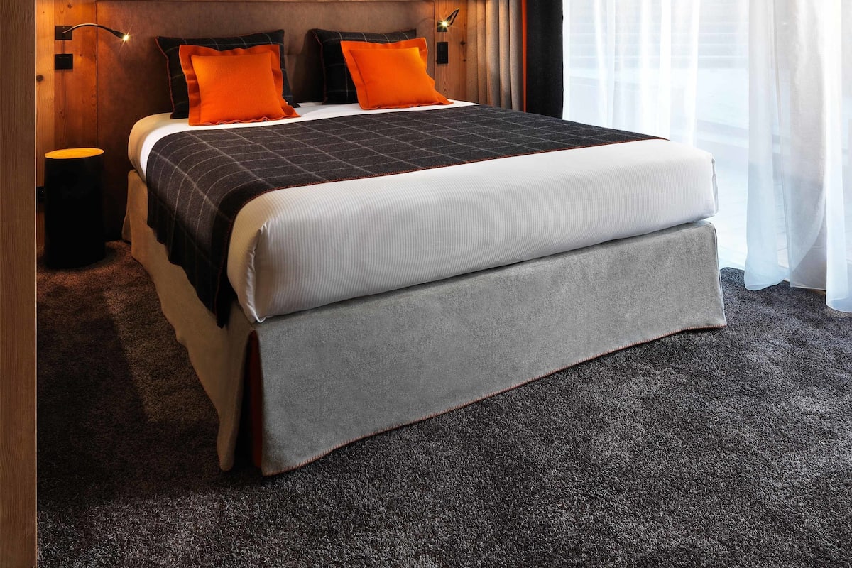 A modern hotel room with a wooden panel wall, a large bed with a grey and white duvet, orange pillows, and two black wall lamps.