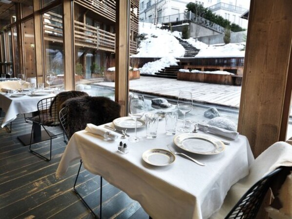 A restaurant with a table set for dining, a fur throw on a chair, and a snowy outdoor view. - Le M de Megève (Megeve)