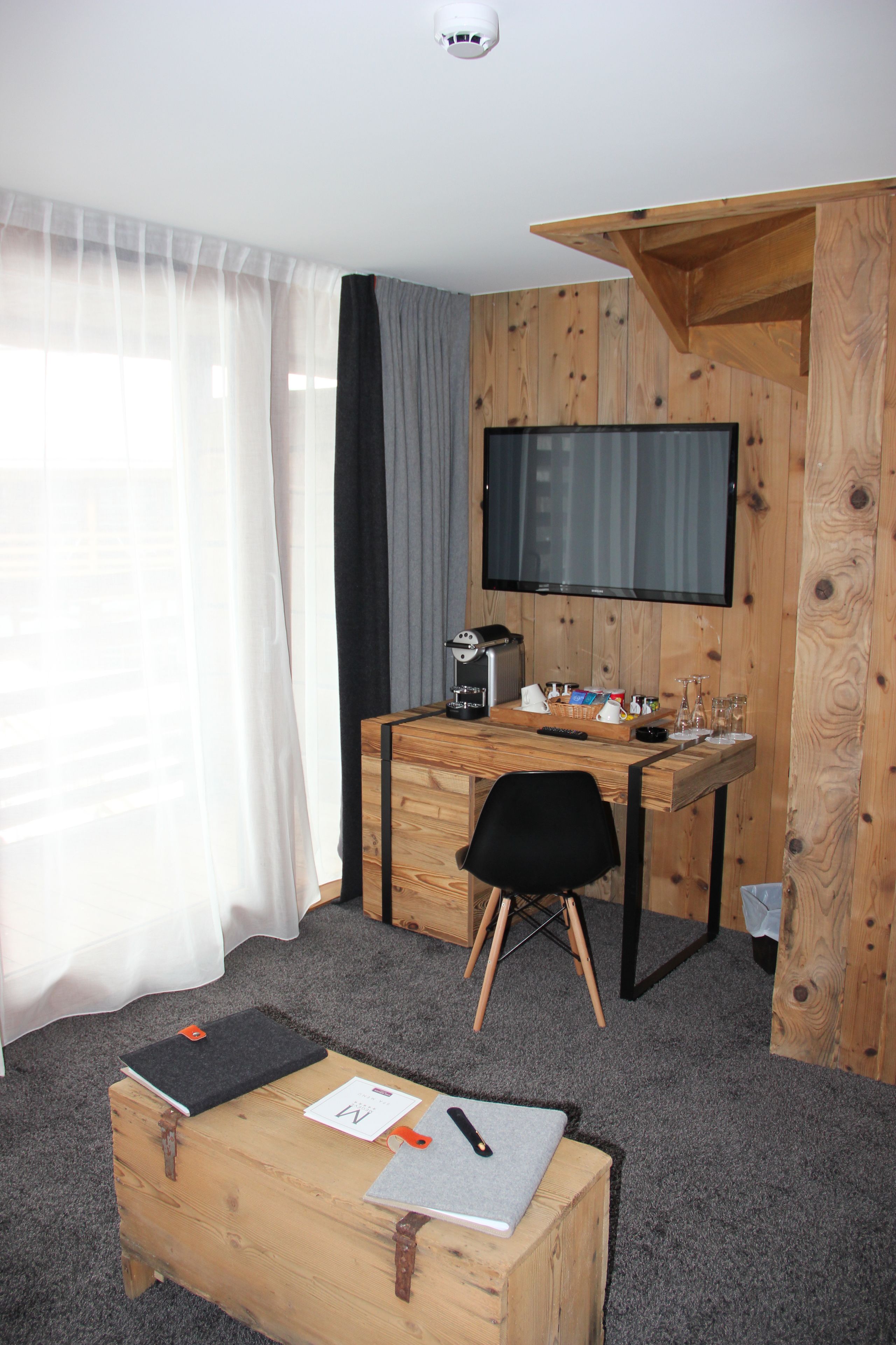 Une pièce de taille modeste, comprenant un bureau en bois, une chaise, un téléviseur fixé au mur et une petite table sur laquelle se trouvent un carnet et un stylo.