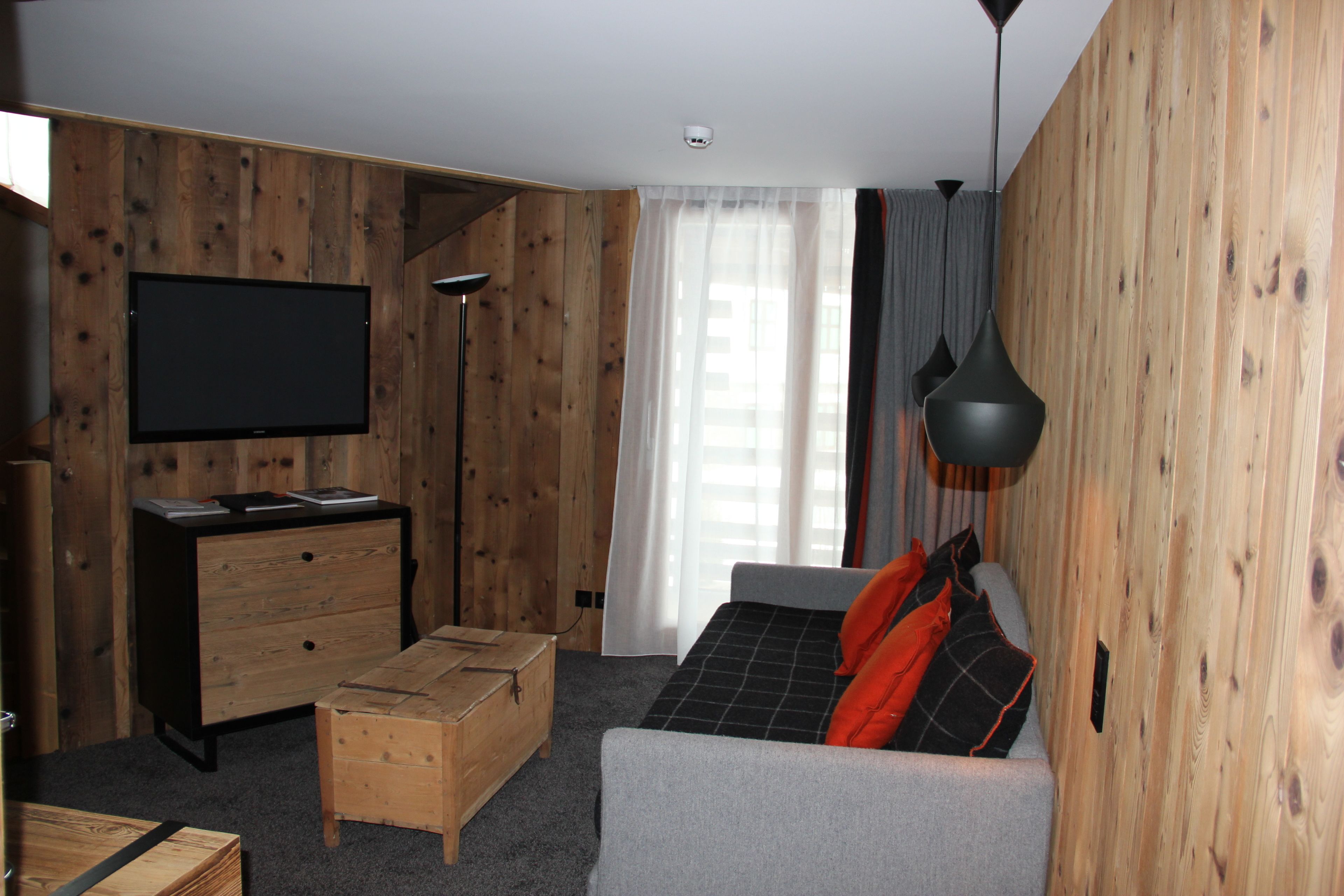A room with a flat-screen TV mounted on the wall, a wooden dresser, a sofa with a black and orange blanket, and a hanging pendant light.